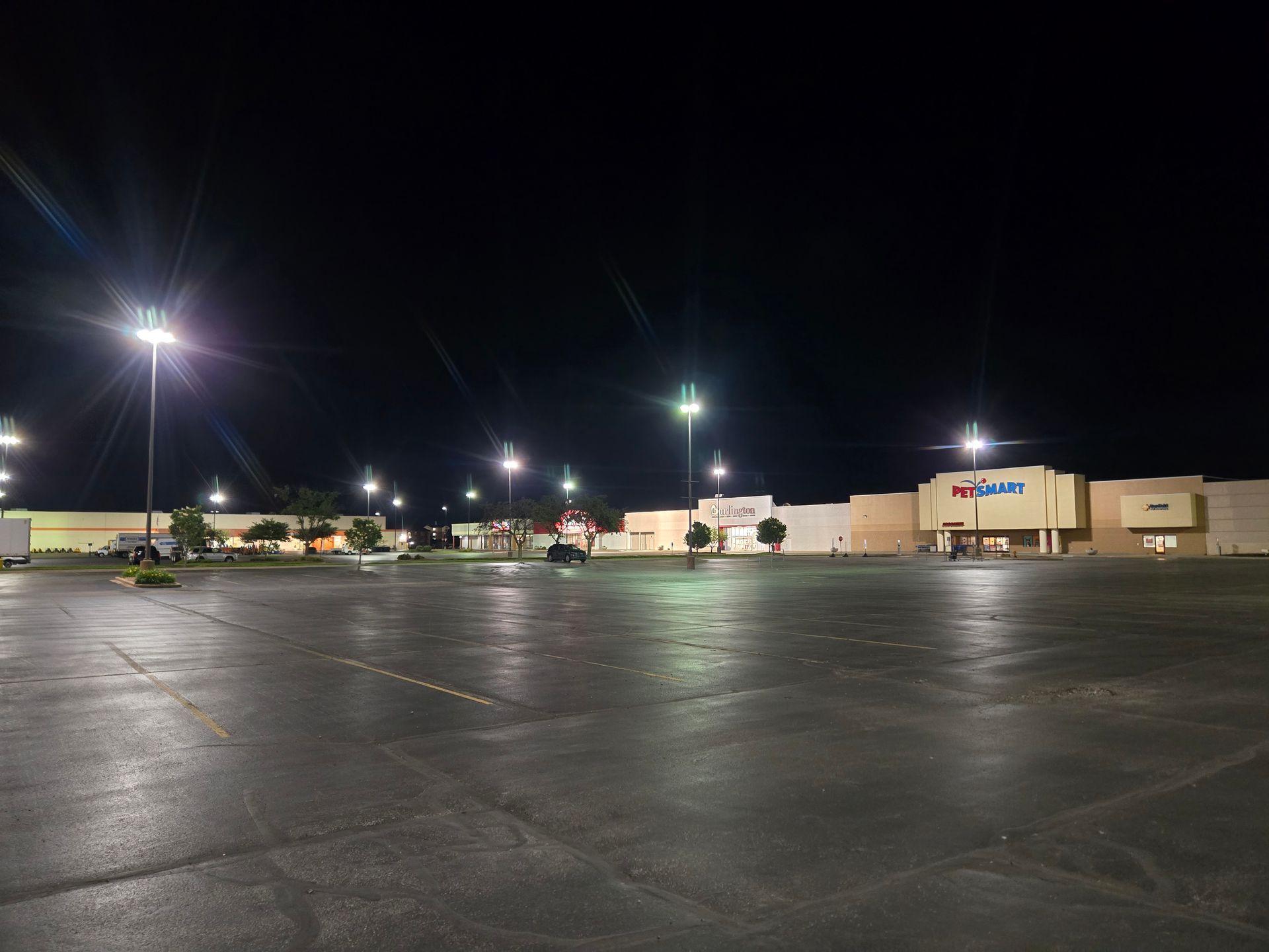 Empty parking lot at night lit by streetlights. Buildings in background.