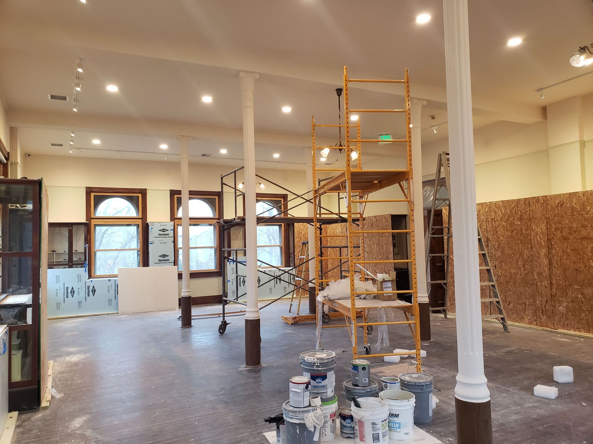 Interior of a room under renovation with scaffolding, paint cans, and exposed walls. Windows and columns are visible.