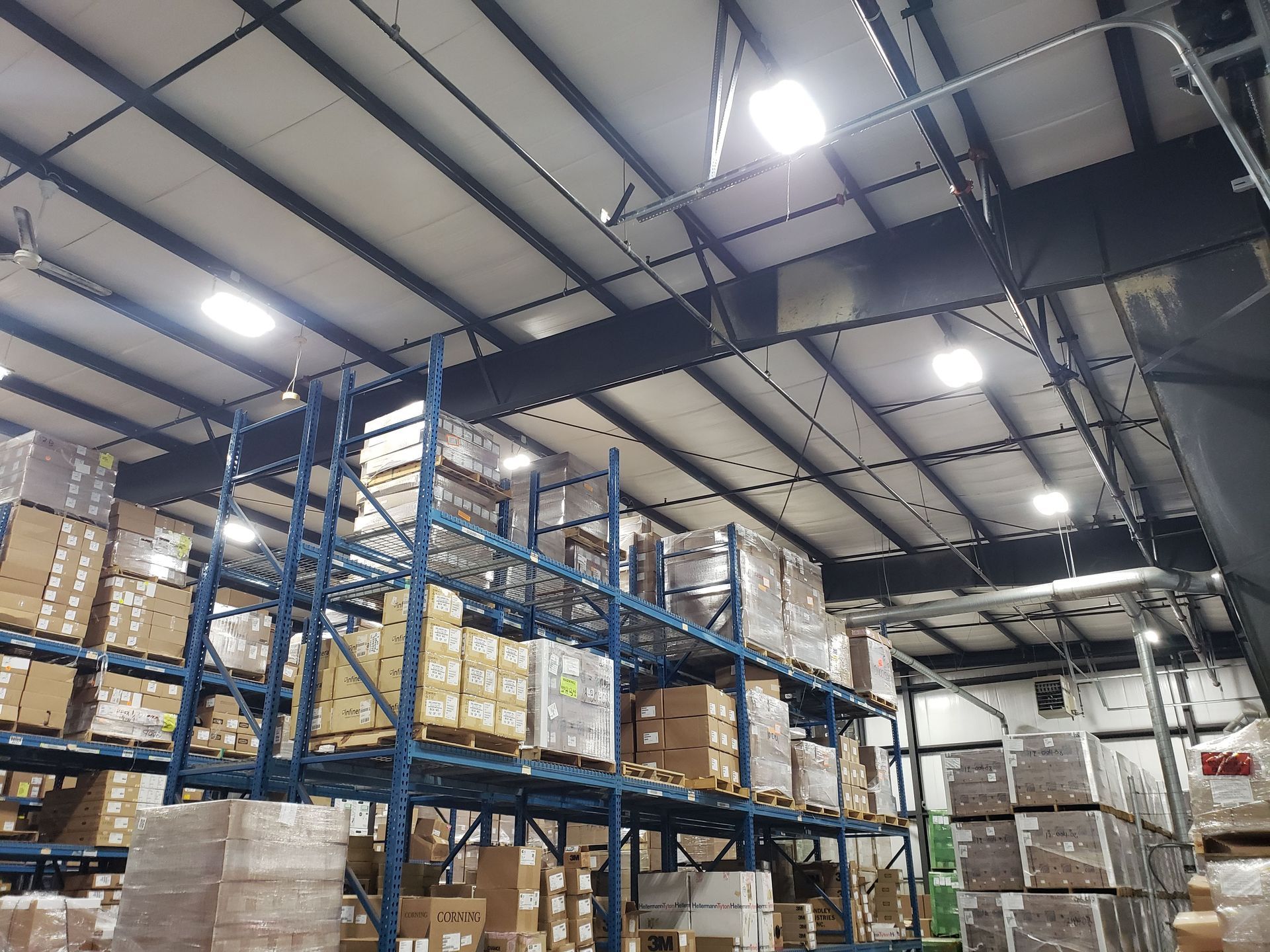 Warehouse interior: blue metal shelving holding stacked cardboard boxes under bright ceiling lights.