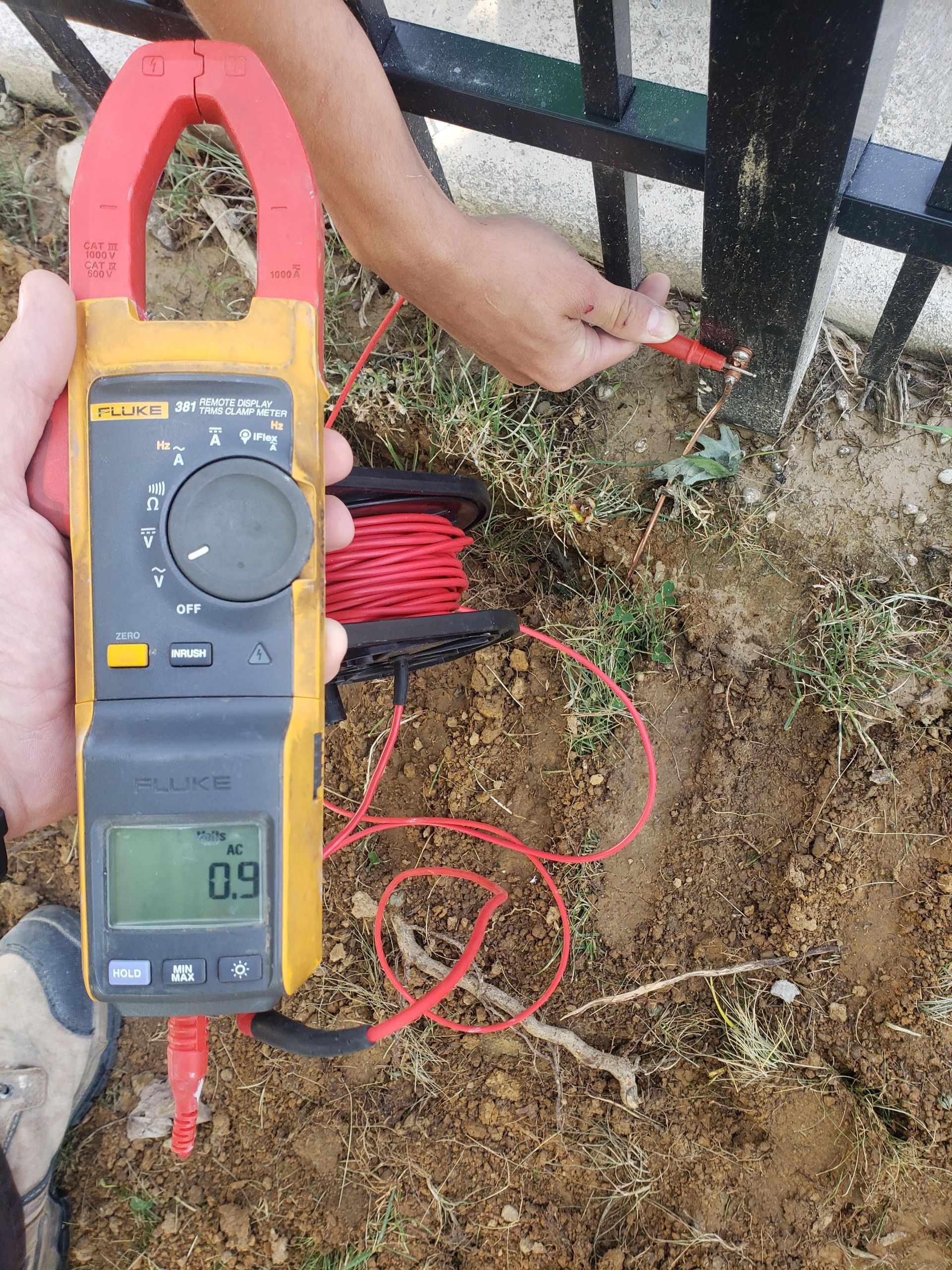A person tests a black metal fence post with a multimeter in an outdoor setting. The meter displays a reading of 0.9.