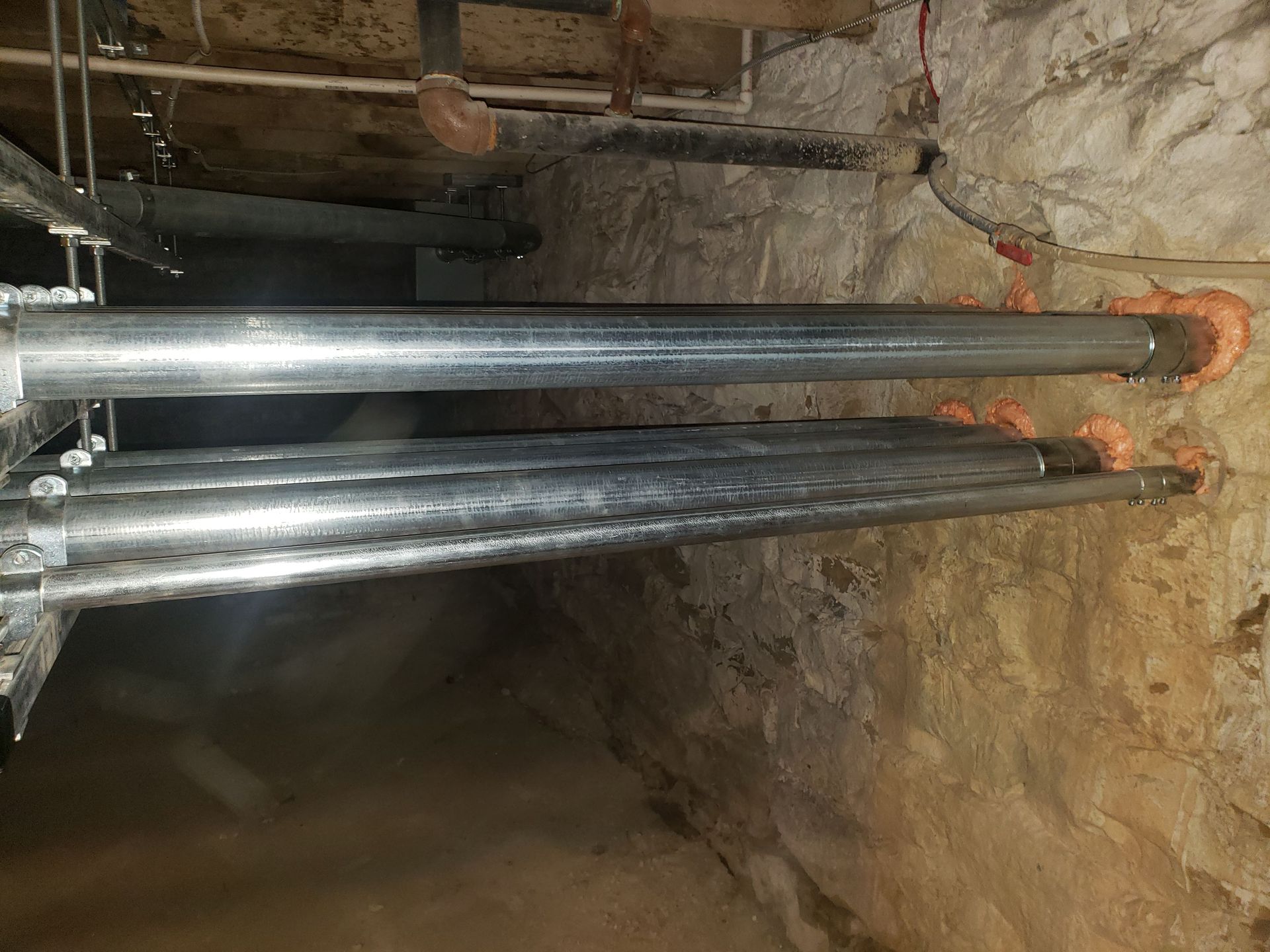 Galvanized pipes running through a stone wall, sealed with orange foam.