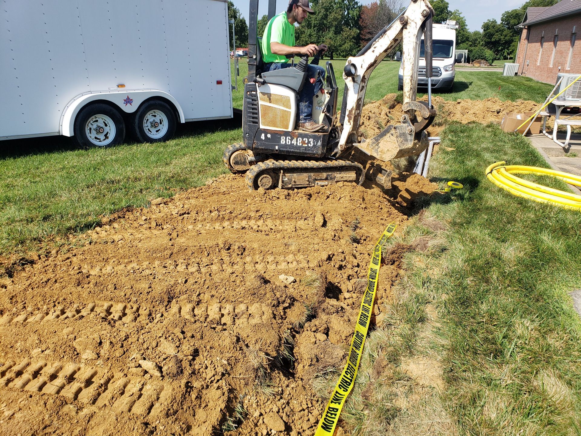 Man operating a Bobcat excavator, digging a trench in a grassy yard. Yellow caution tape and a trailer are visible.