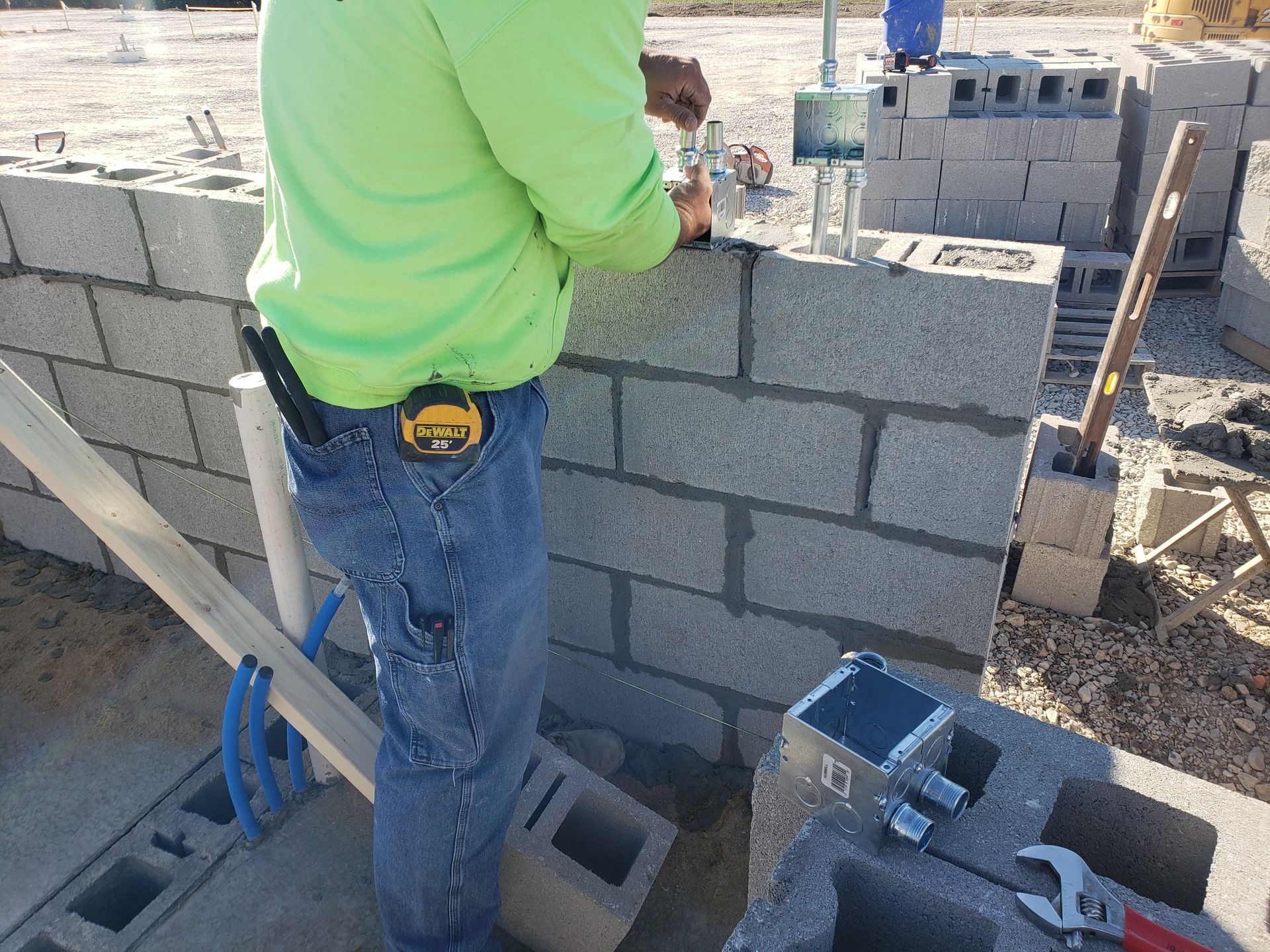 Construction worker installing electrical box on cinder block wall. Bright green shirt, blue jeans, outdoor setting.