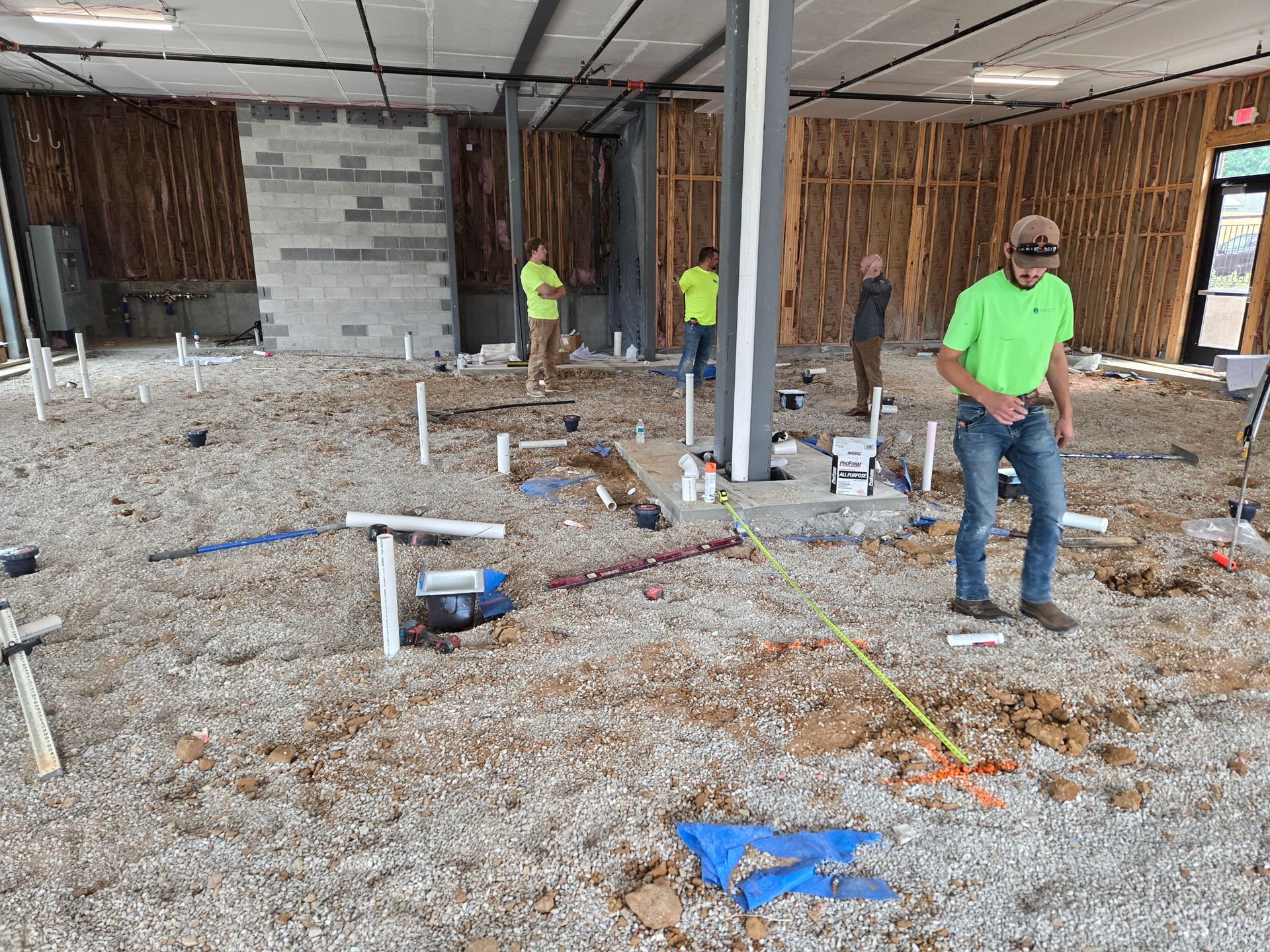 Construction site interior with workers; exposed plumbing, gravel floor, unfinished walls, and ceiling.