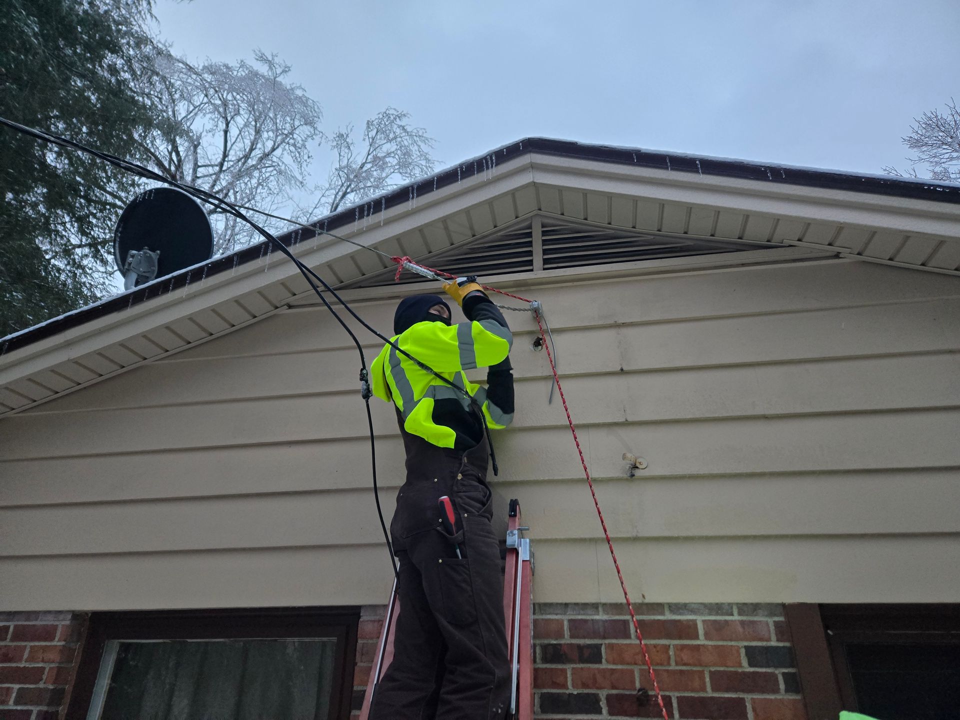Person in reflective gear on a ladder fixing electrical wires on a house, overcast day.