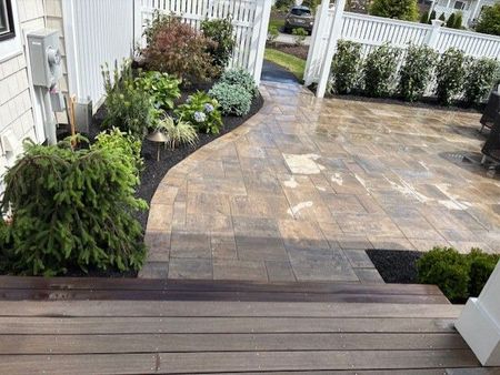 A paved patio and pathway lead past landscaped beds toward a white fence. The pathway's edge curves, and a set of wooden steps lead down.