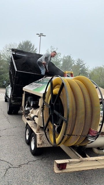 A man dumping material from a truck bed into a large yellow hose reel on a trailer, outdoors in a parking lot.