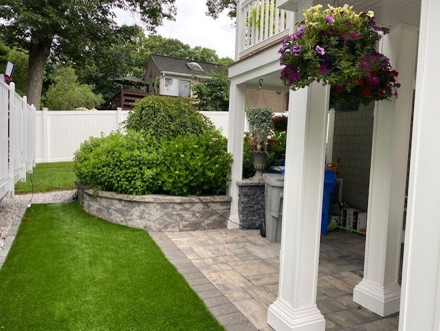 A backyard patio with green turf and a white fence. Features a flowering hanging basket, stone wall, and white columns.