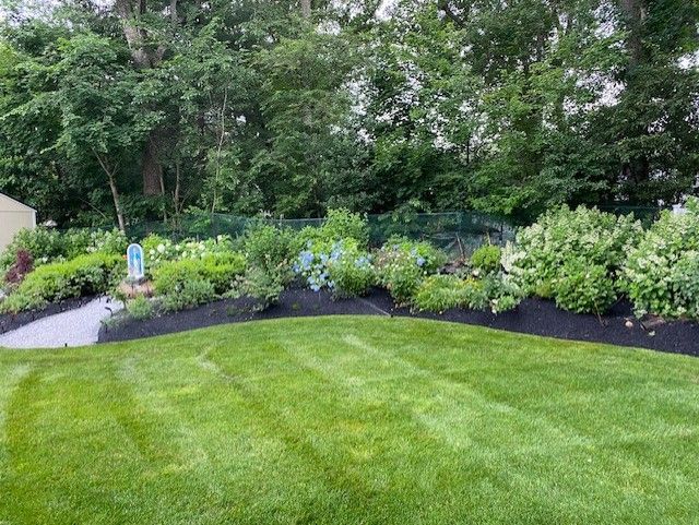 A lush green lawn leads to a flower bed with various plants and a backdrop of trees. The flowerbed is mulched.