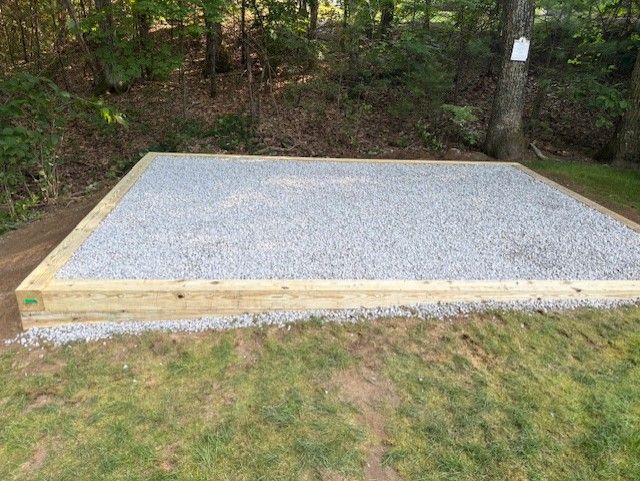 Gravel-filled rectangular base, likely for a shed, framed by wooden beams on a grassy lawn. Forest in the background.