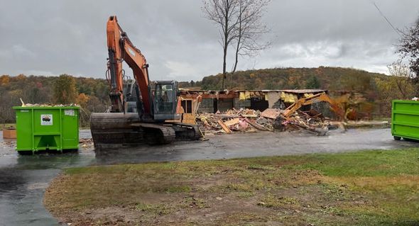 An excavator demolishes a building, debris piled in front. Green dumpsters sit nearby. Overcast sky, fall foliage.