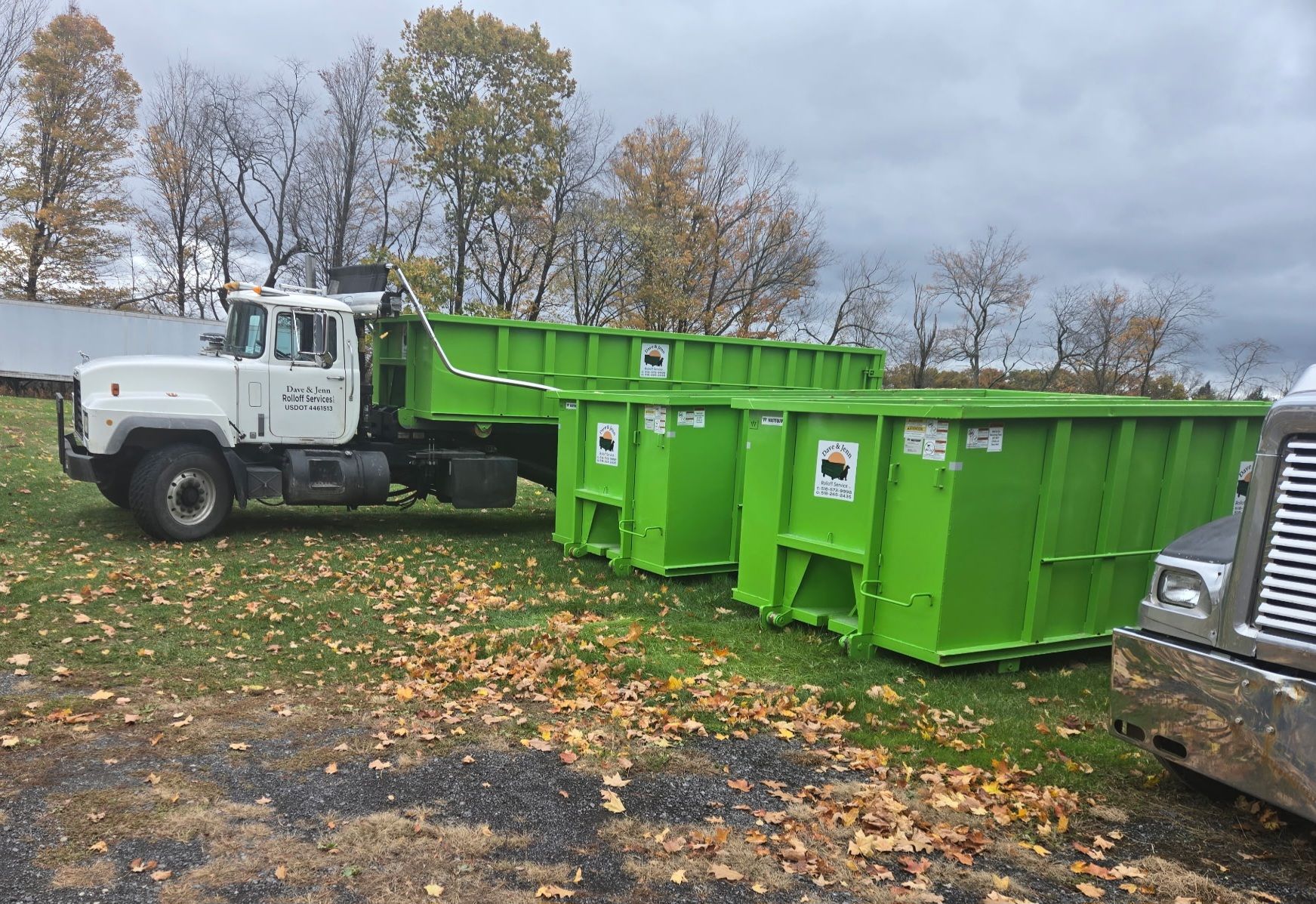 Green dumpsters next to a white truck on a grassy area, likely for waste disposal.