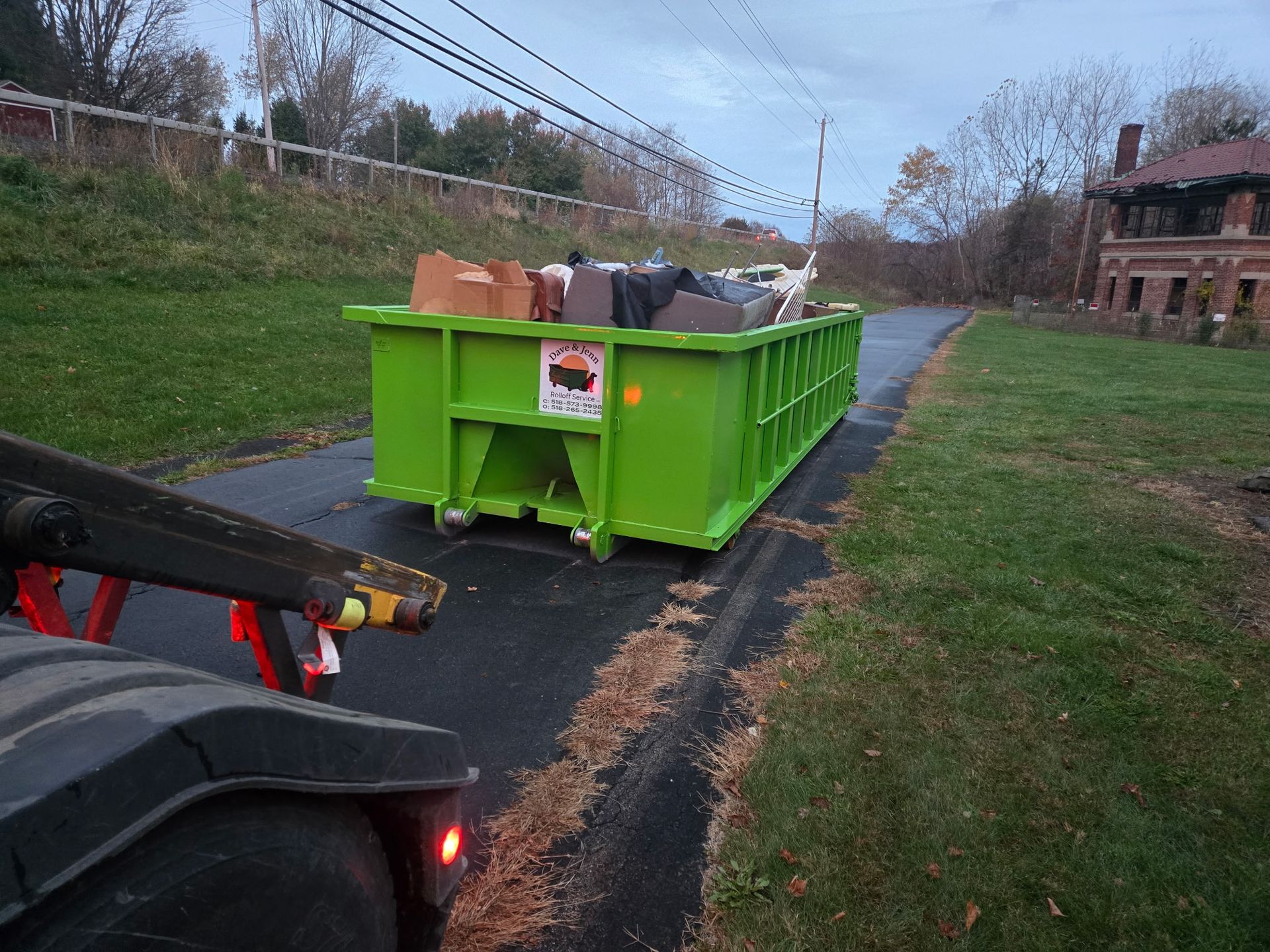Green dumpster on a paved road, filled with trash, being towed by a truck.