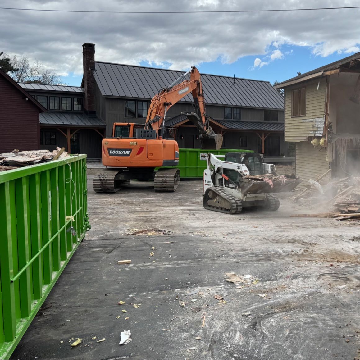 Demolition site with excavator tearing down a building, skid steer moving debris, green dumpsters.