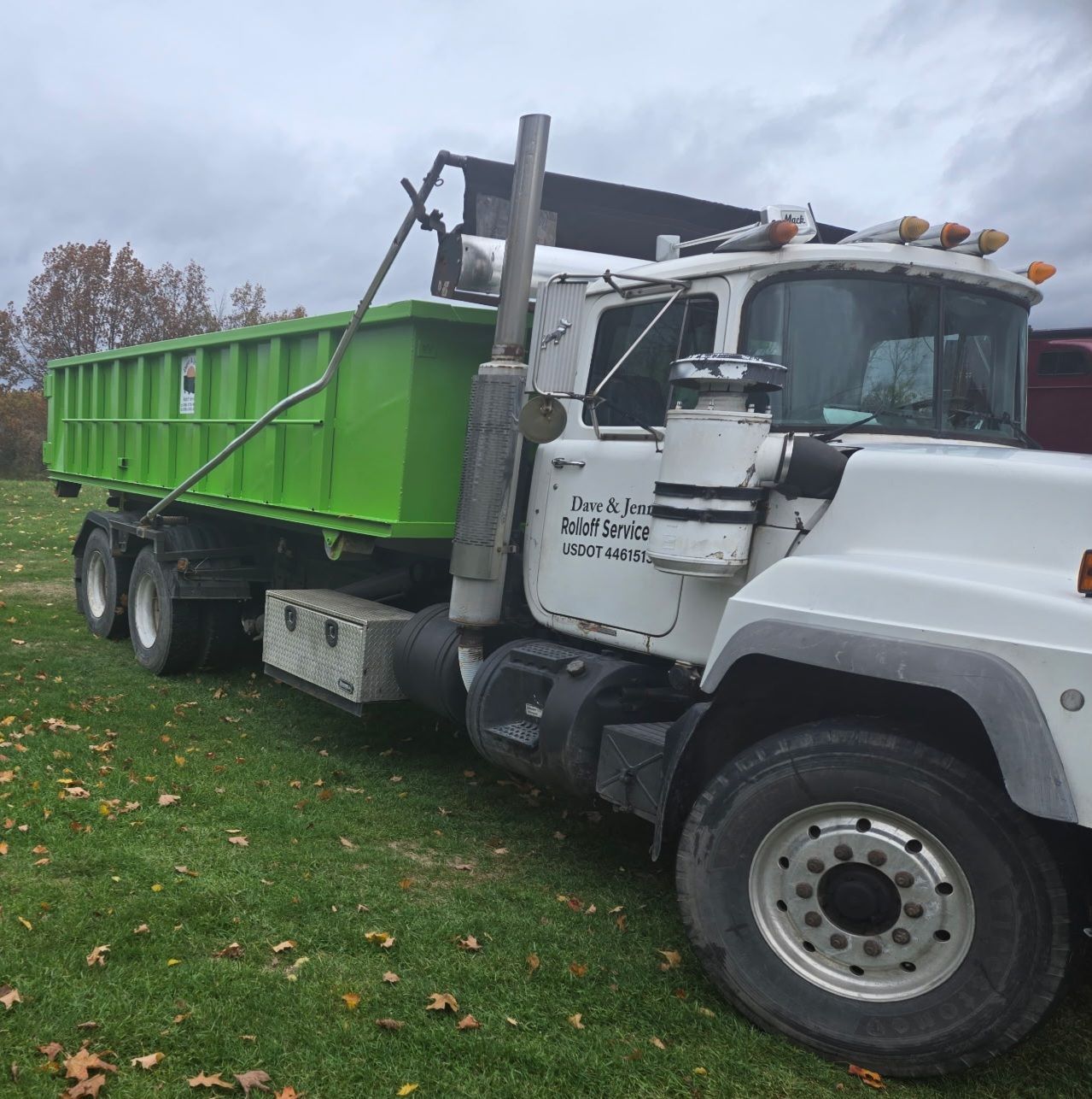 White Mack roll-off truck with a green dumpster on a grassy area under an overcast sky.