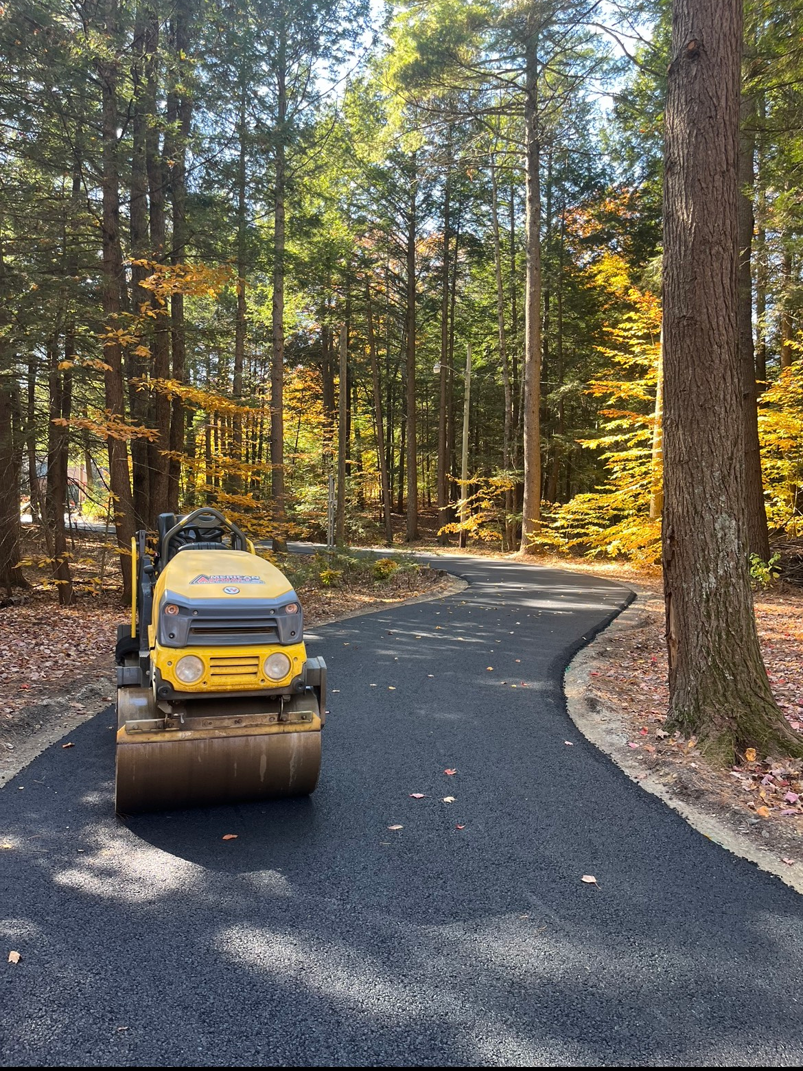 A yellow roller is driving down a road in the woods.
