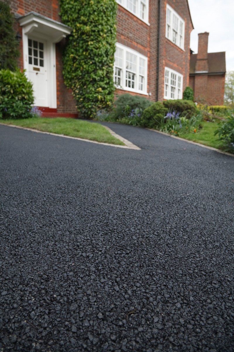 Dark gray driveway leading to a brick house with a white front door and climbing greenery.