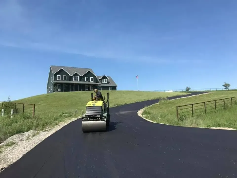 Asphalt roller compacting a newly paved driveway leading to a green house on a hill.