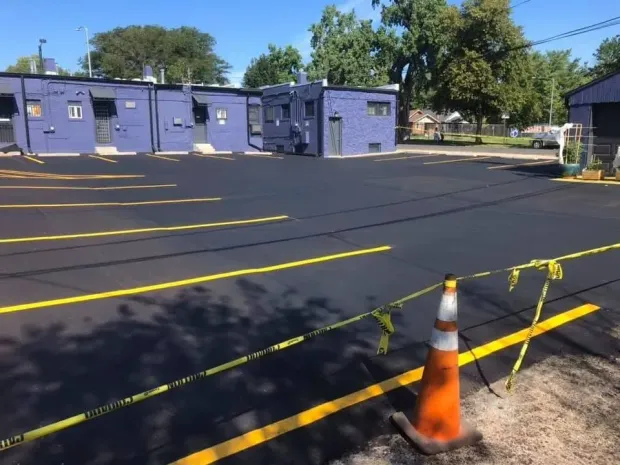 Newly paved parking lot with yellow lines and blue buildings. Orange cone with caution tape in foreground.