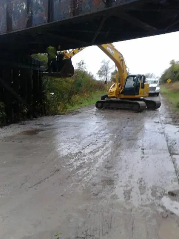 A yellow excavator is driving under a bridge on a muddy road.