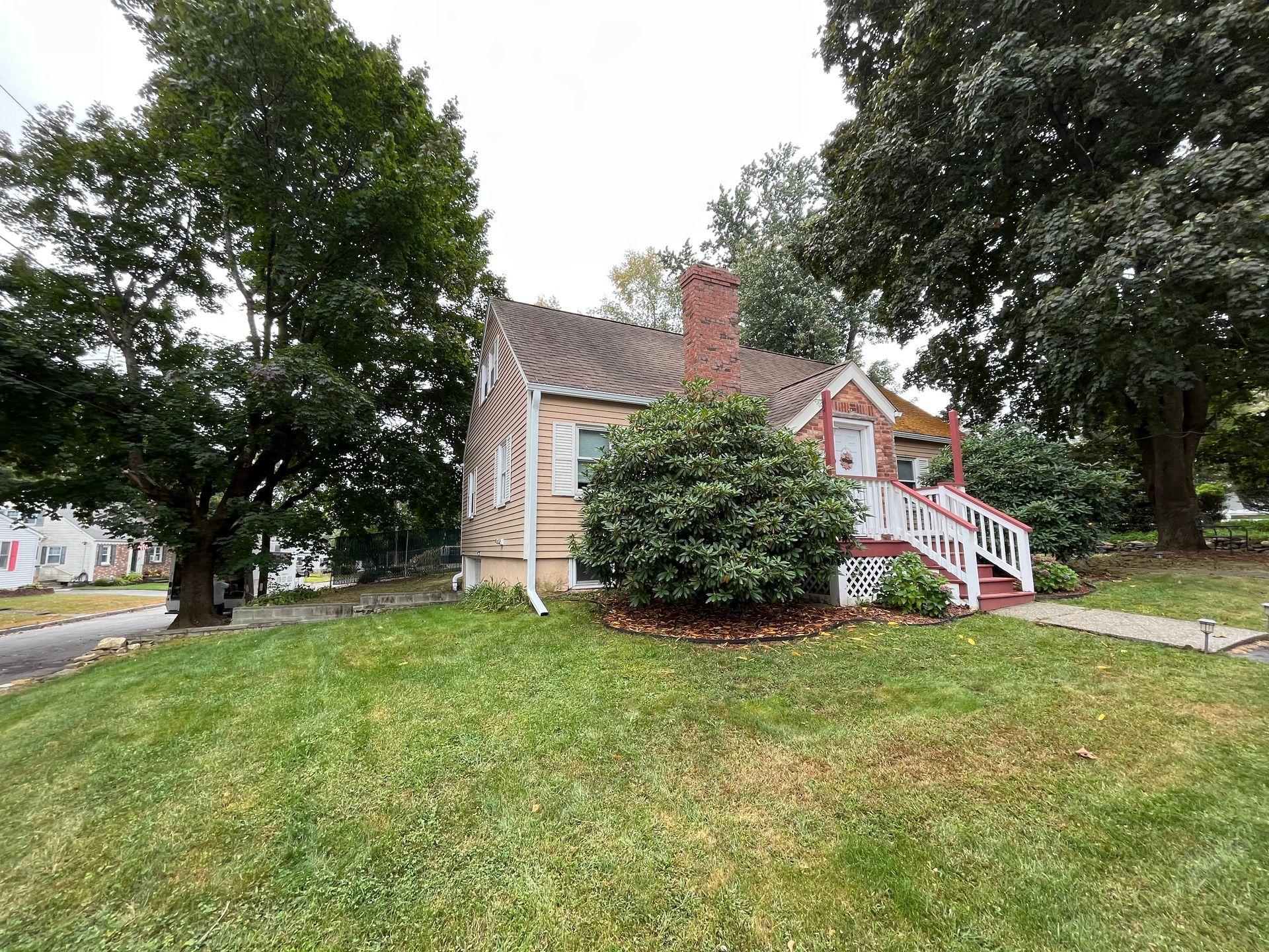 Small beige house with red stairs and chimney on a grassy lot, framed by large trees