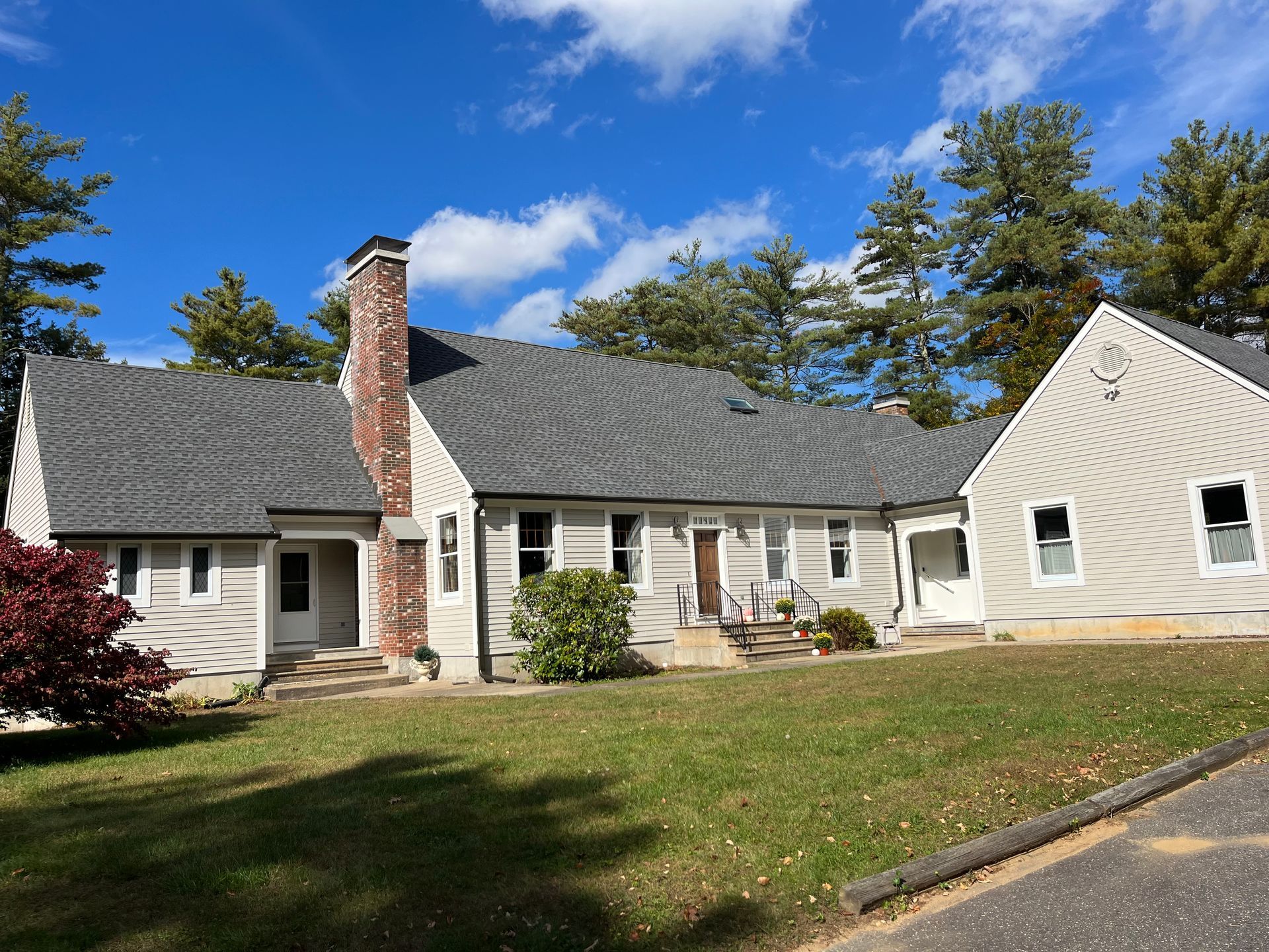 Single-story beige house with gray roof, brick chimney, and a grassy front yard under blue sky.