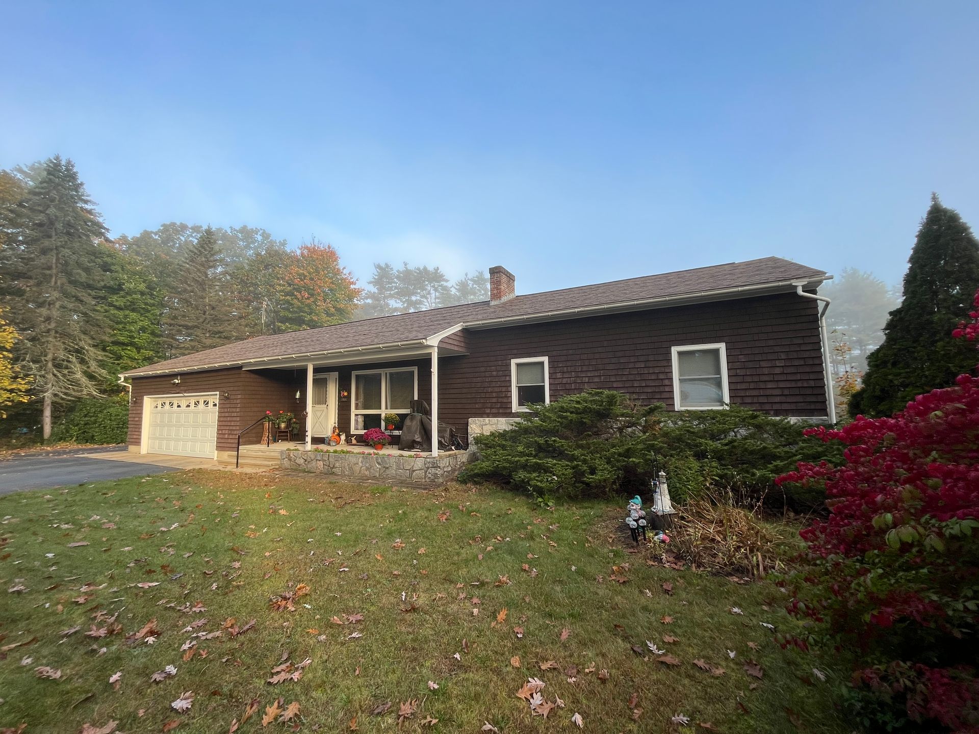 Single-story house with stone porch, brown siding, and a grassy yard with autumn leaves