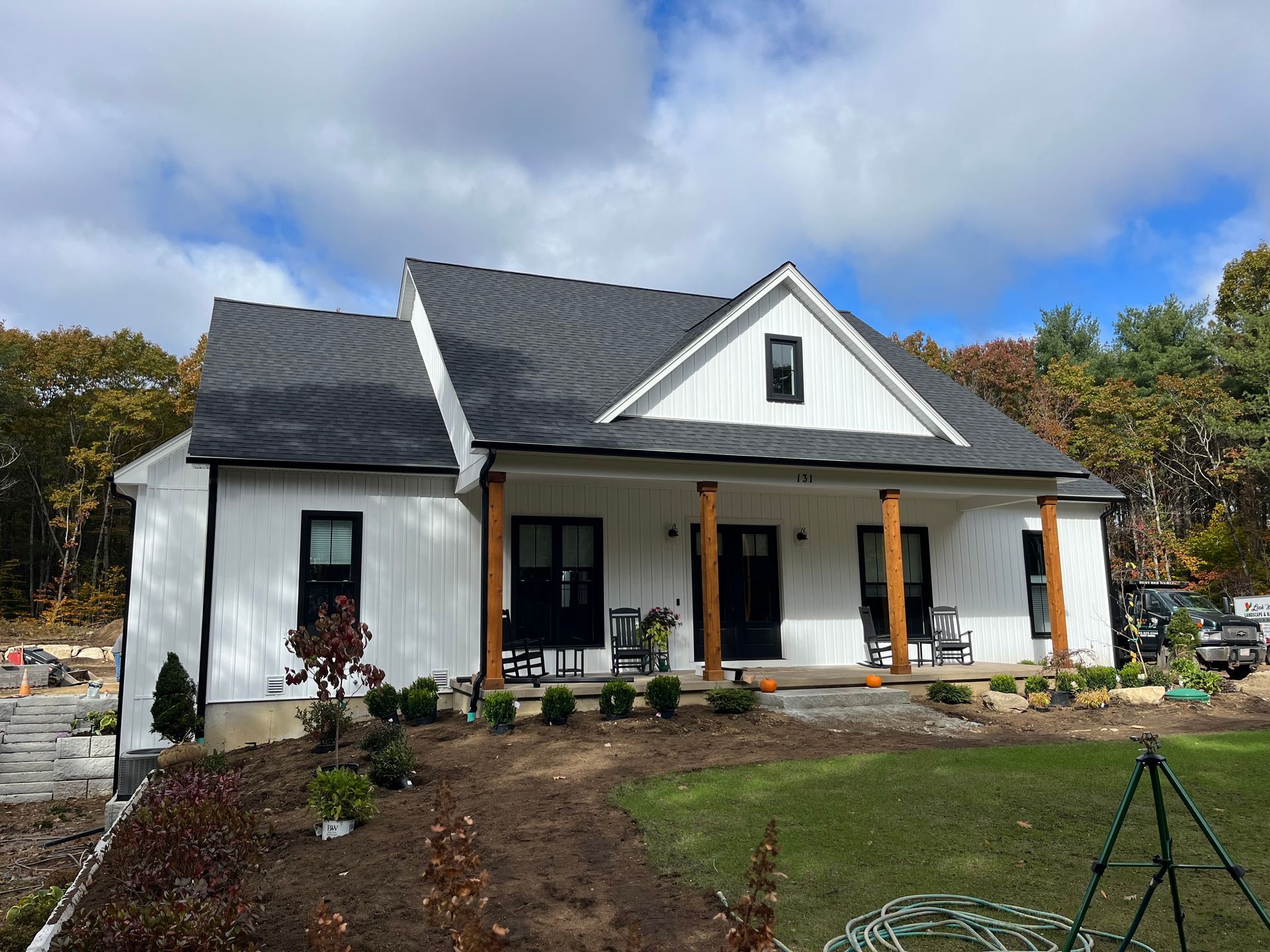 White cottage with black roof and porch in a landscaped yard under a cloudy sky