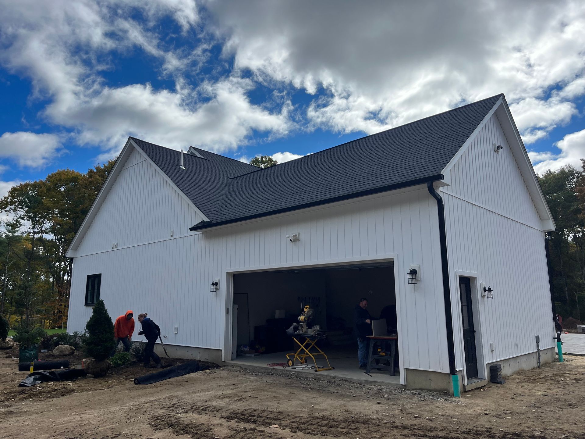 White barn-style garage with open door, black roof, and two people working outside under a cloudy sky