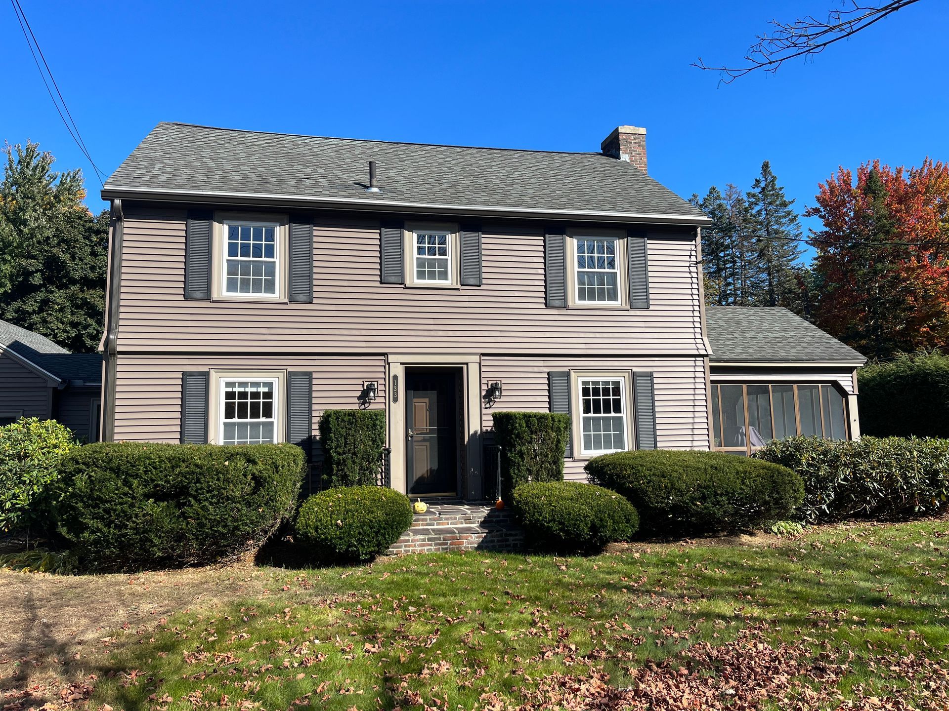 Two-story beige house with white shutters, a front porch, and trimmed bushes under a clear blue sky