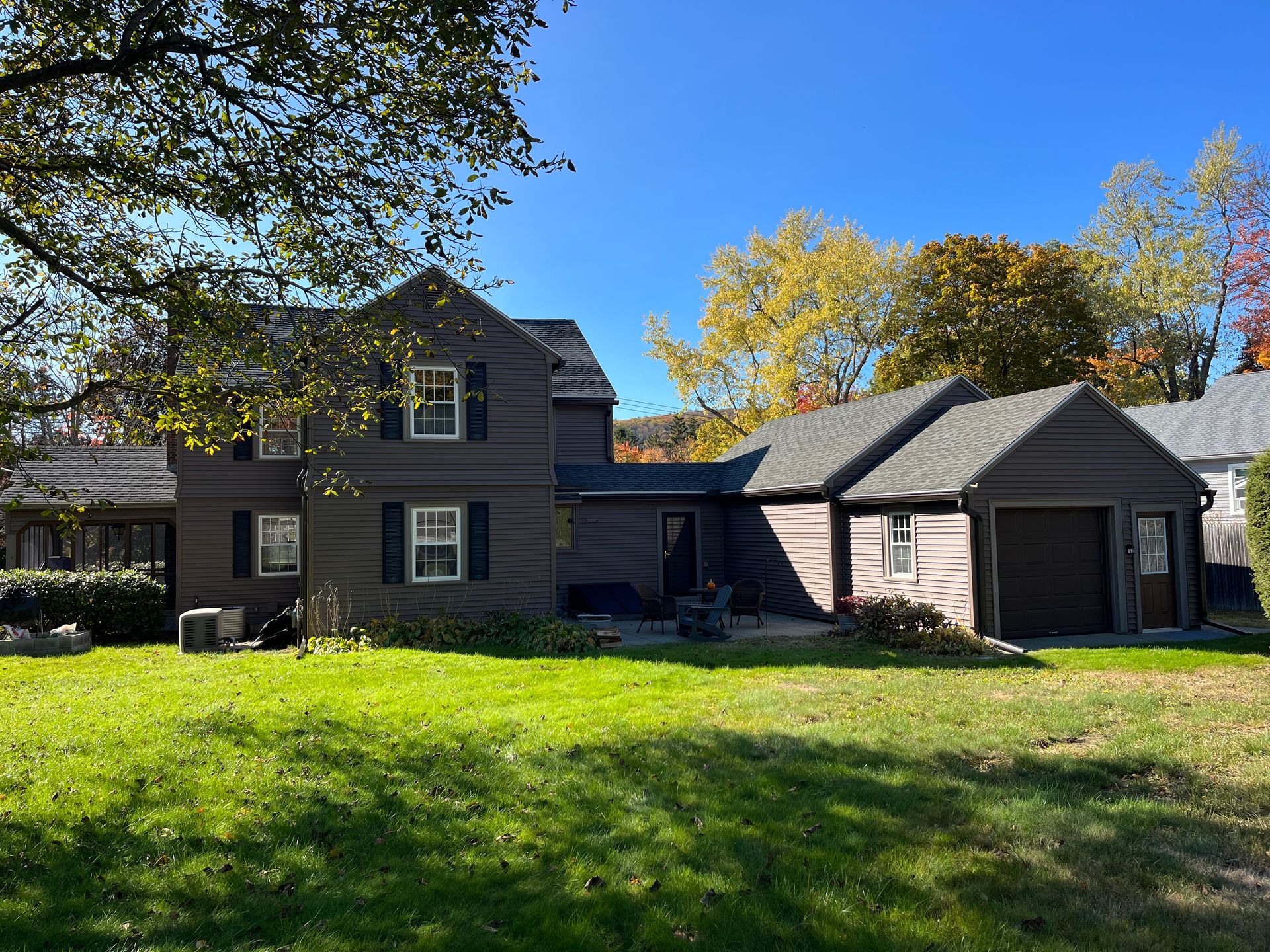 Suburban house with gray siding, attached garage, and a green lawn under sunny autumn trees