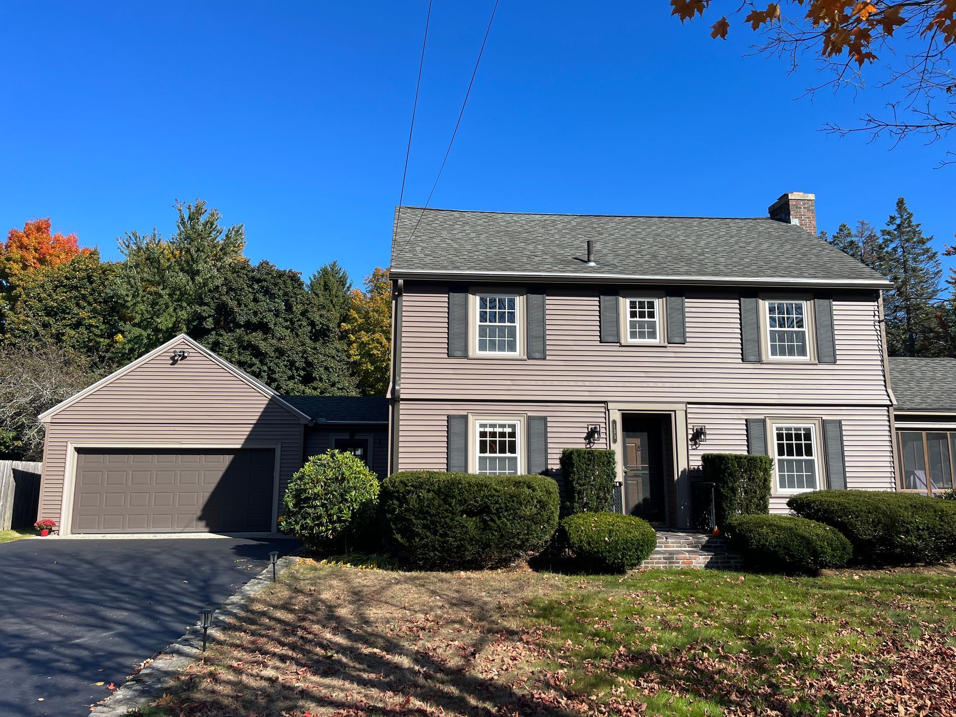 Two-story beige house with detached garage, dark roof, and landscaped front yard under a clear blue sky