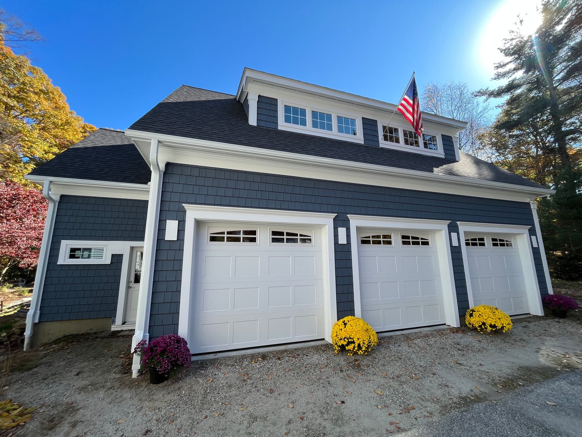 Blue-gray garage with white double doors, flower pots, and an American flag under a bright sky.