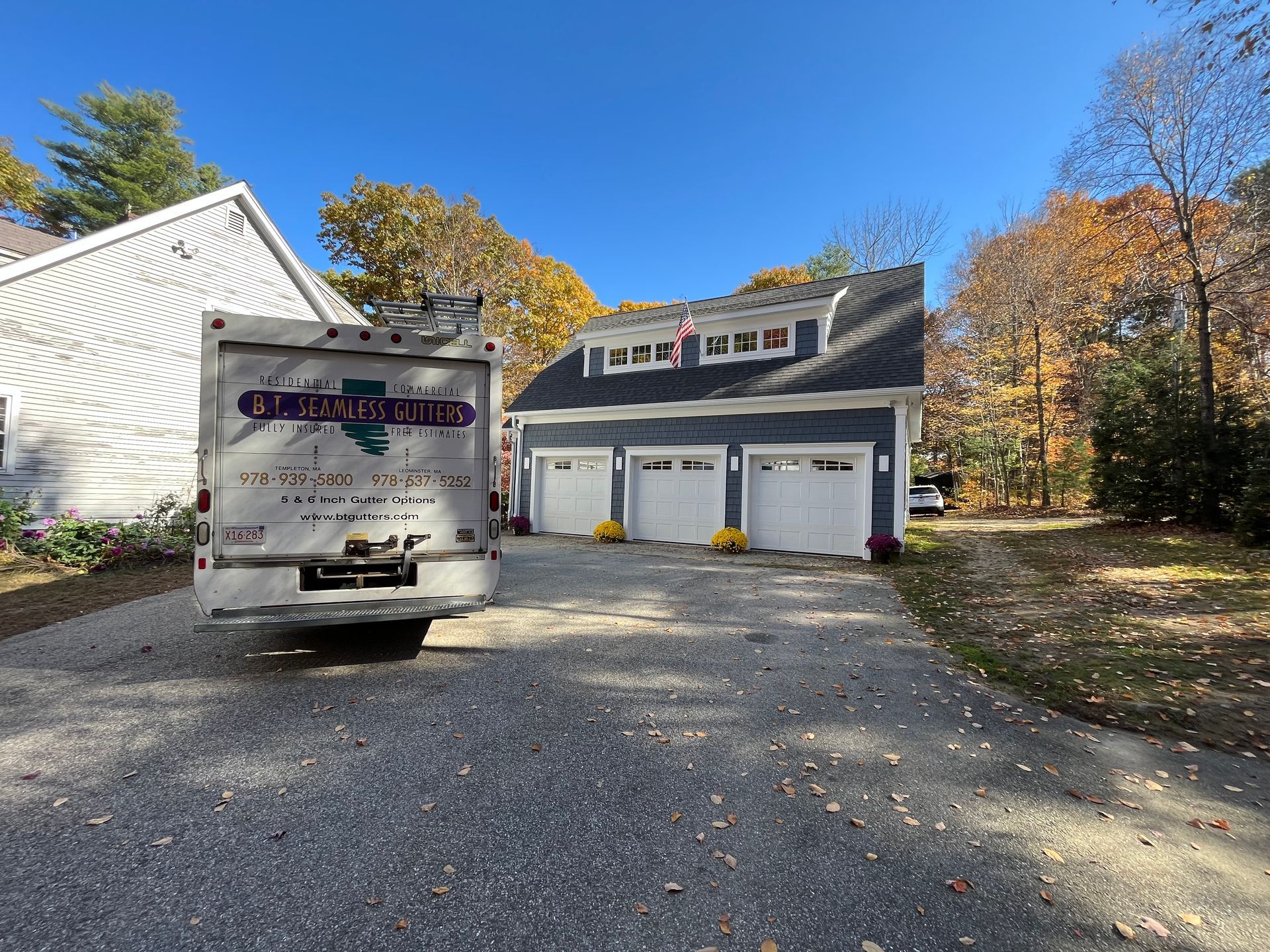 Driveway leading to a three-car garage with a moving truck parked in front, surrounded by autumn trees.