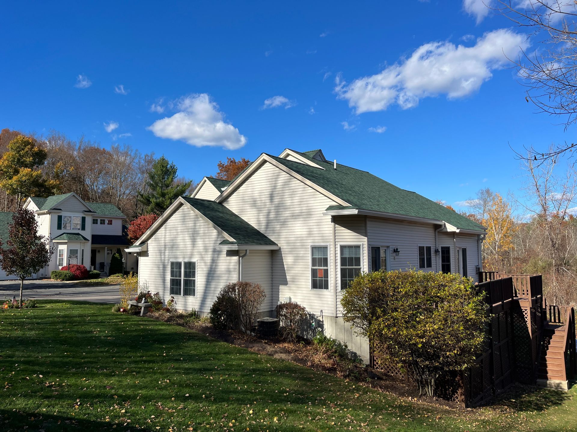 White house with green roof in a sunny yard under a blue sky, with trees and bushes around it