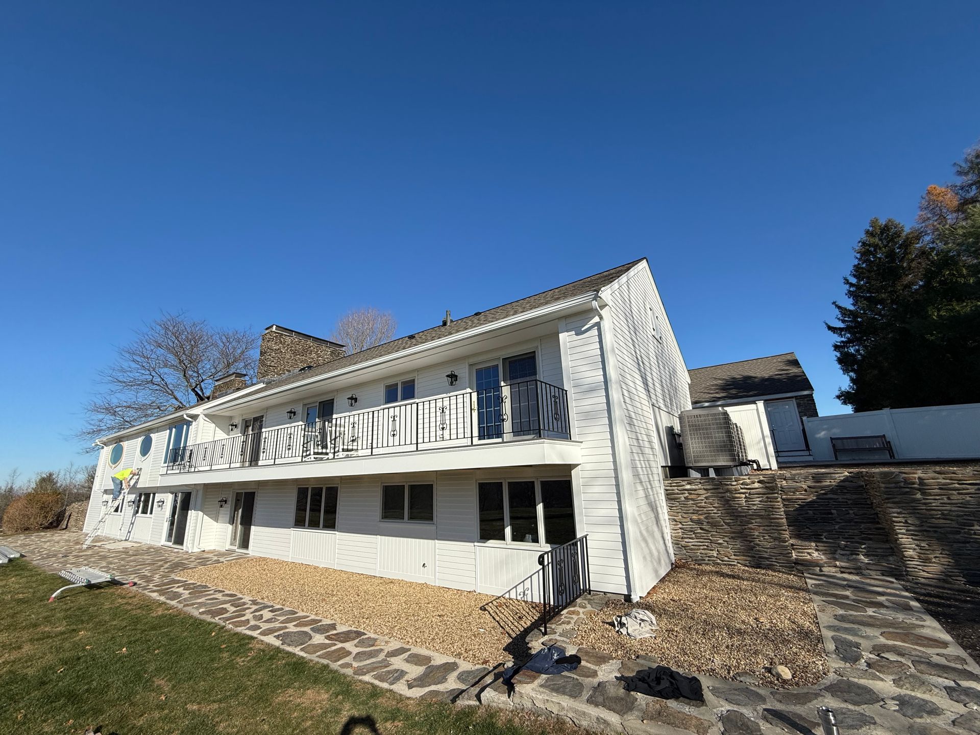 White two-story house with balconies and stone steps on a sunny hillside under a clear blue sky