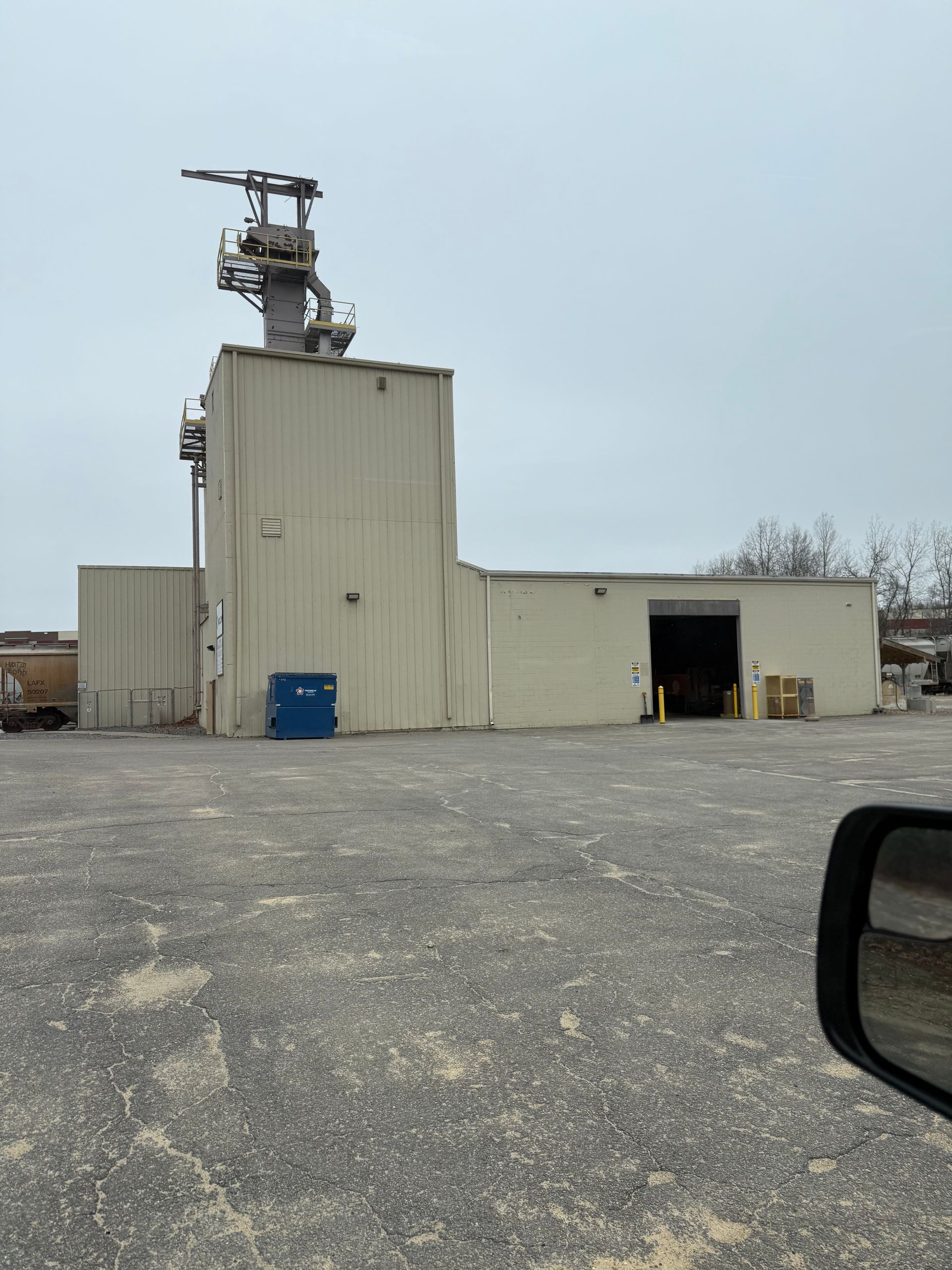 Industrial building with rooftop equipment, a blue dumpster, and a large gravel lot under overcast skies