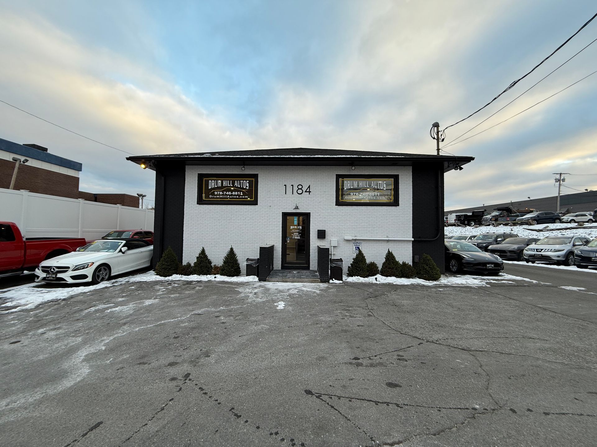 Snowy parking lot in front of a small white building with black trim and signs under a cloudy sky.