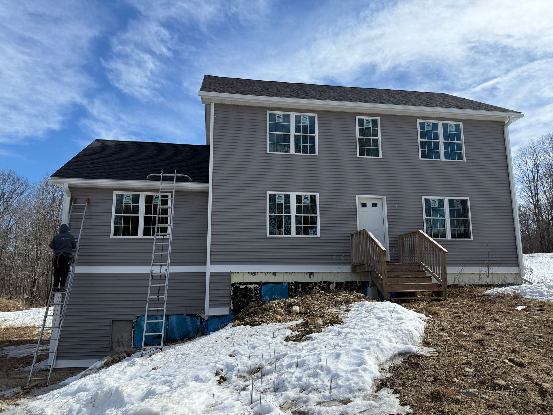 Gray house under construction with white trim, snow-covered yard, and a ladder against the left side
