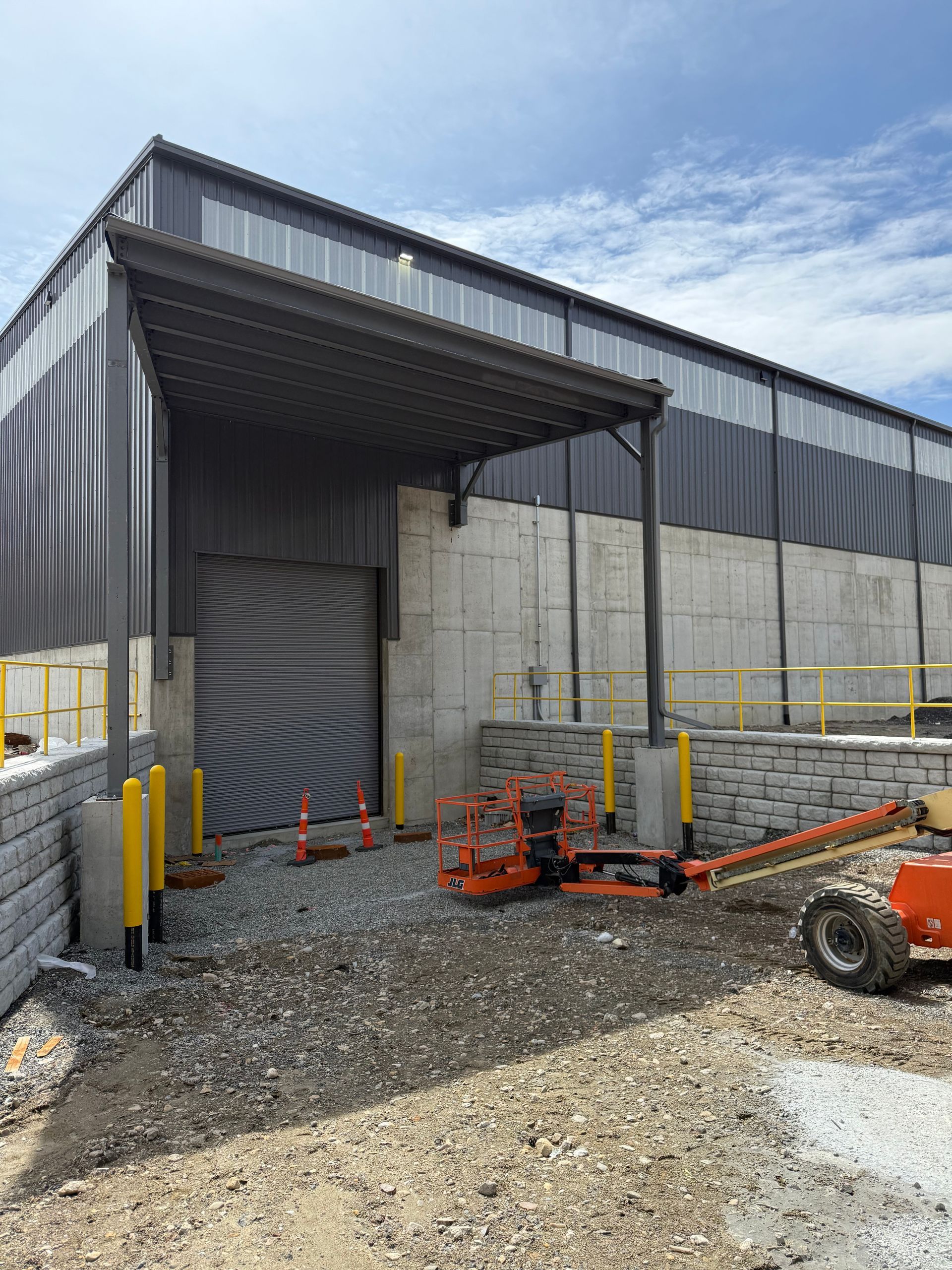 Industrial warehouse exterior with metal siding, concrete walls, and construction equipment on gravel.