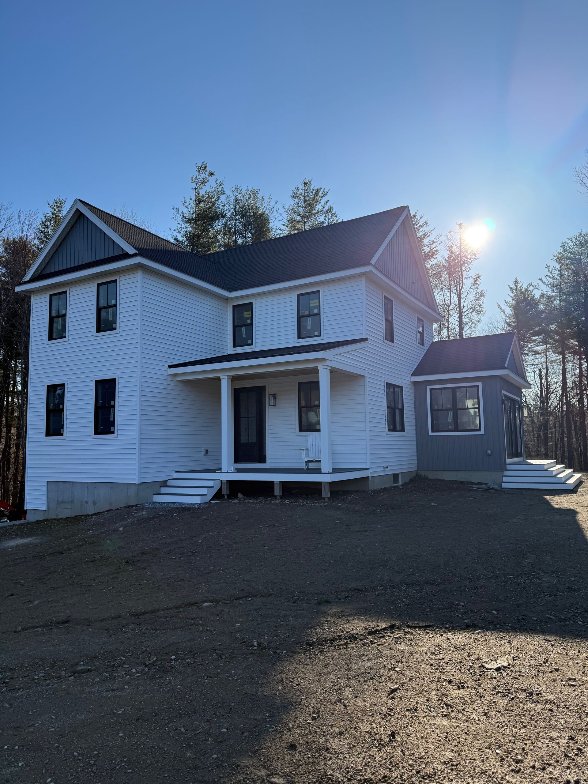 White two-story house with a front porch and attached side addition, lit by bright sun on a gravel driveway.