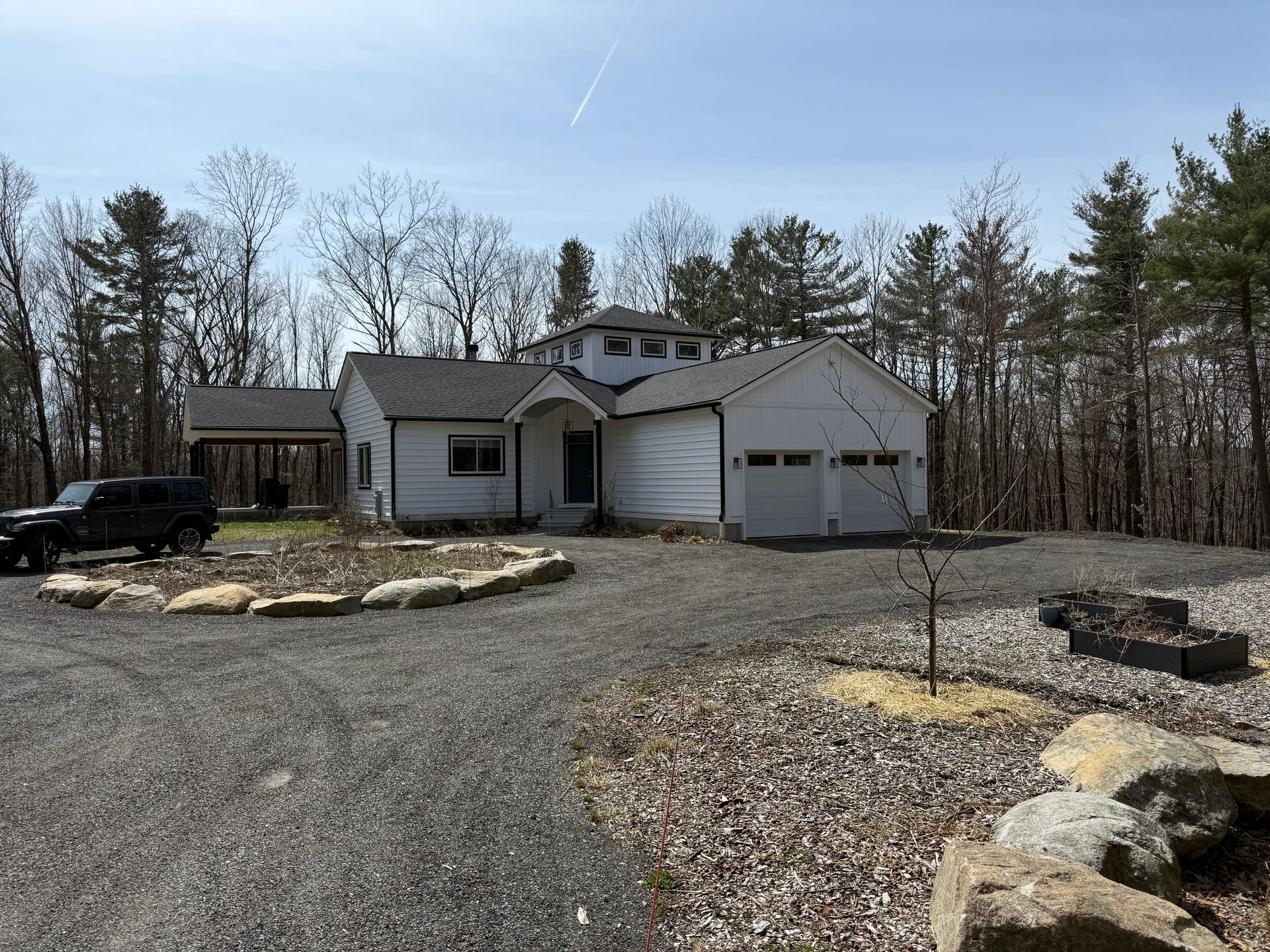 Gravel driveway leads to a white house and garage beside a wooded lot under a clear sky.