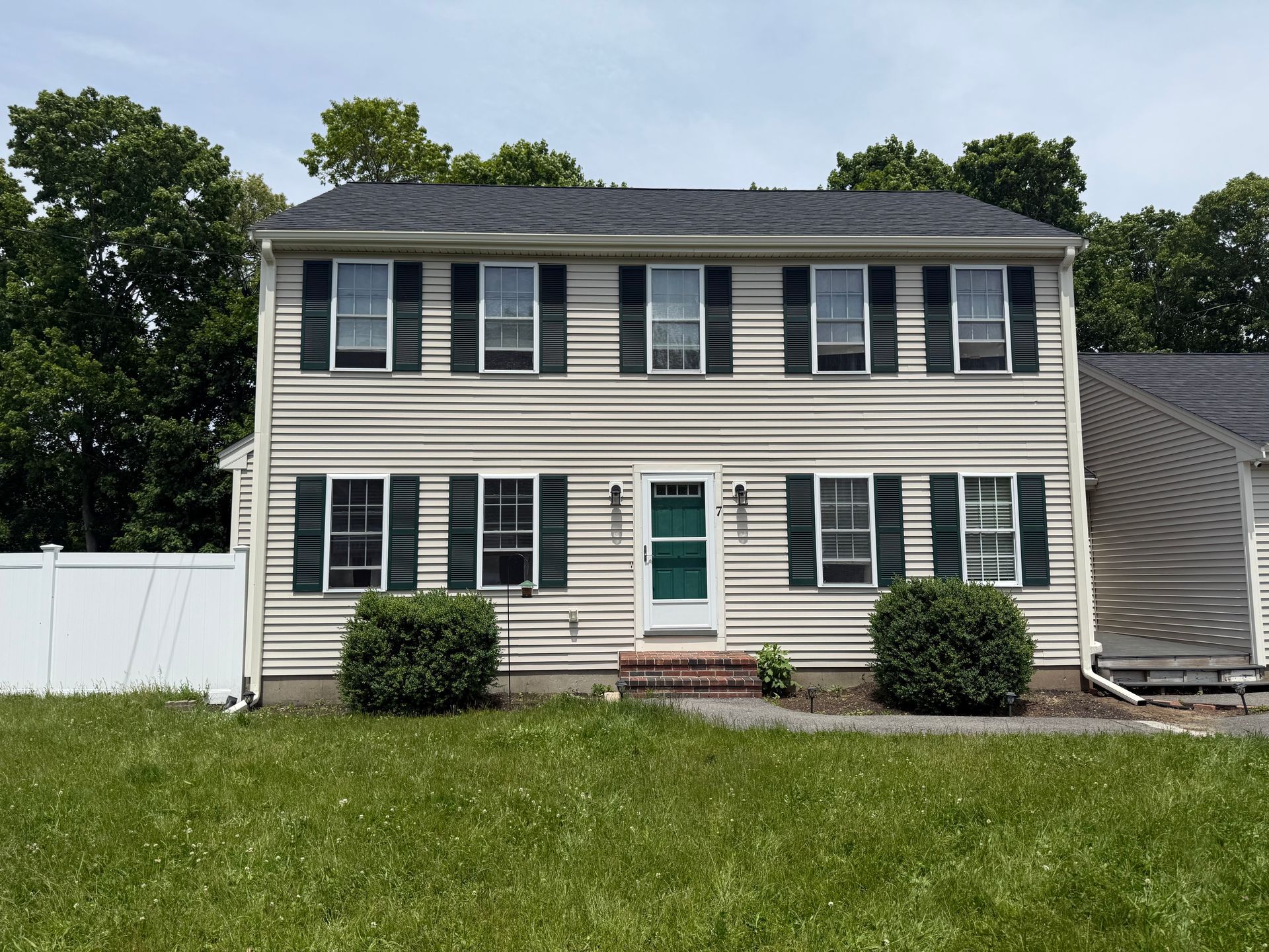 Two-story beige house with dark shutters, white fence, and a green front door, viewed from the front lawn