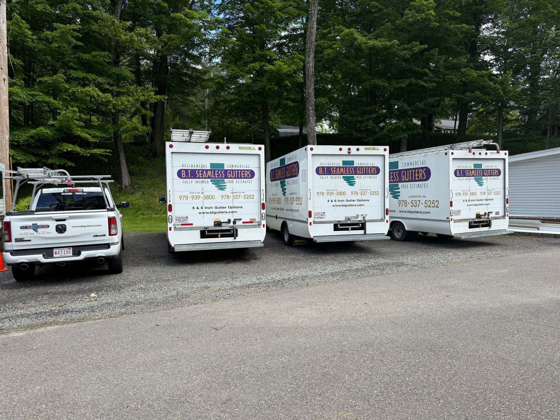 White utility trucks parked in a gravel lot beside trees, one pickup on the left