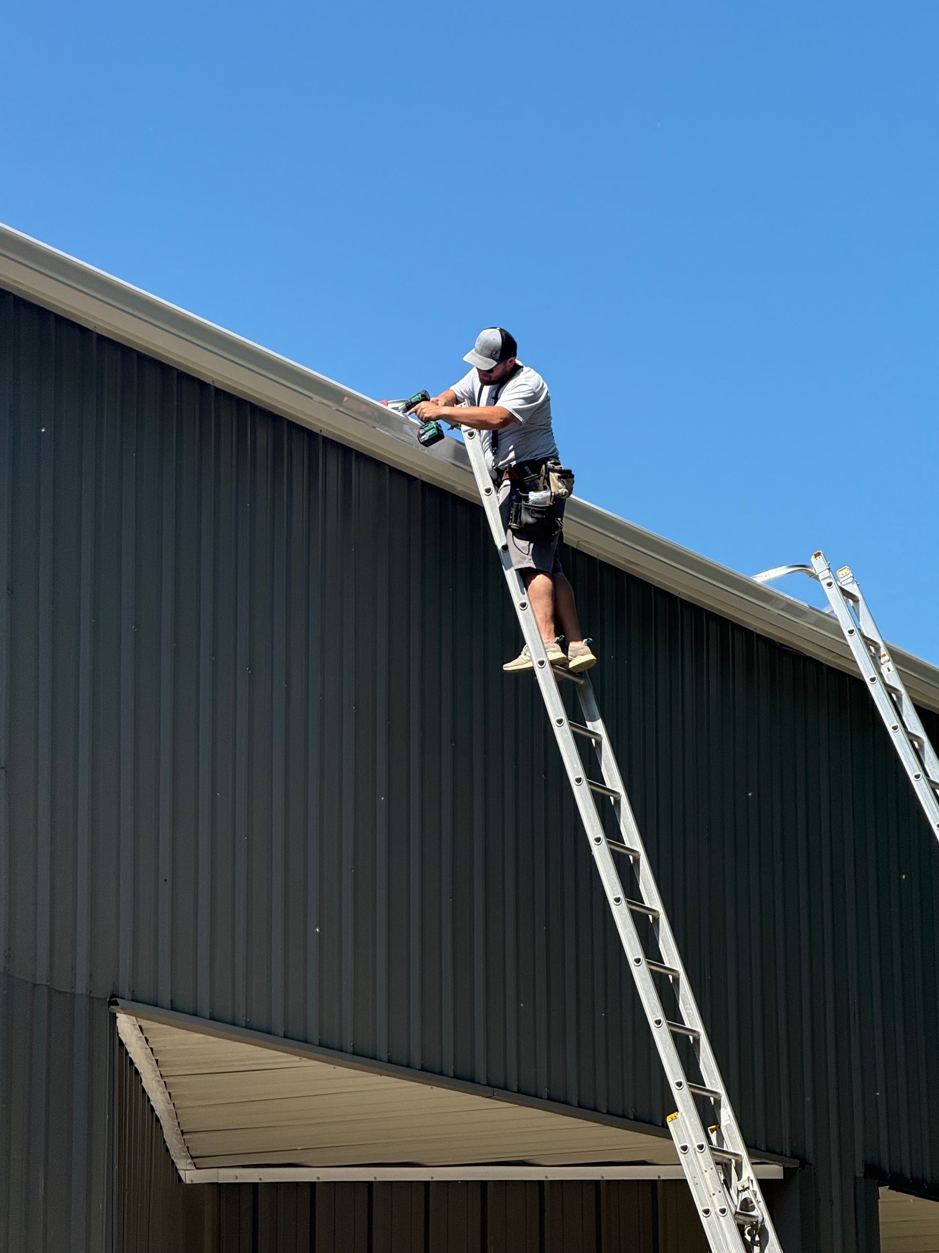 Worker on a ladder inspecting the roof edge of a dark metal building under a clear blue sky