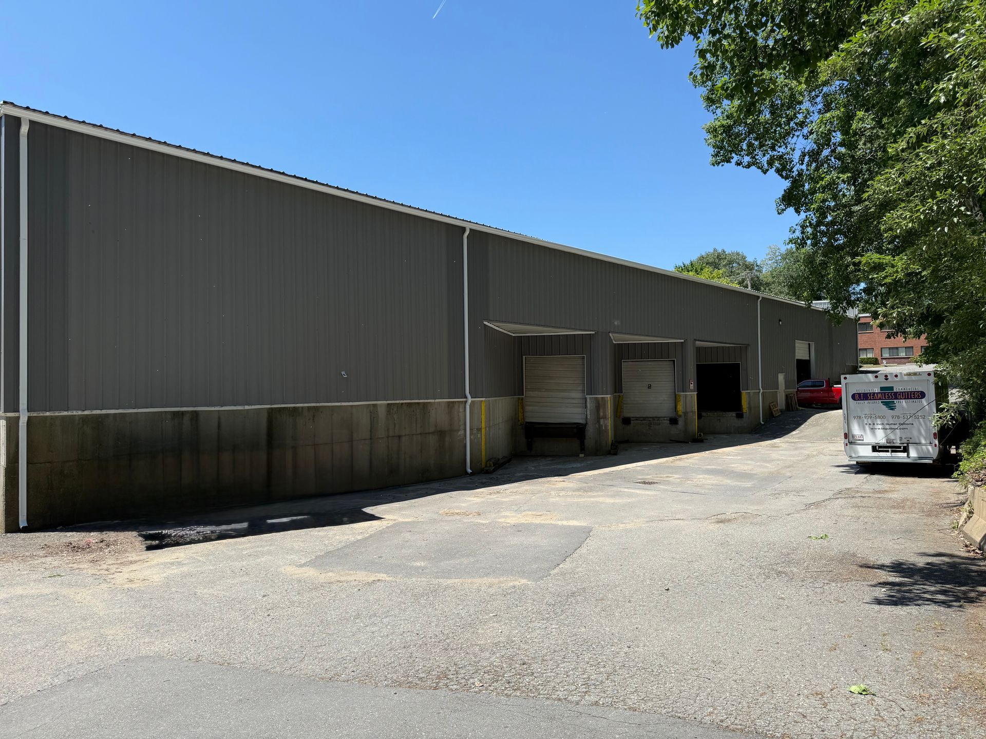 Industrial warehouse loading docks beside a paved lot, with a white truck and trees under a blue sky