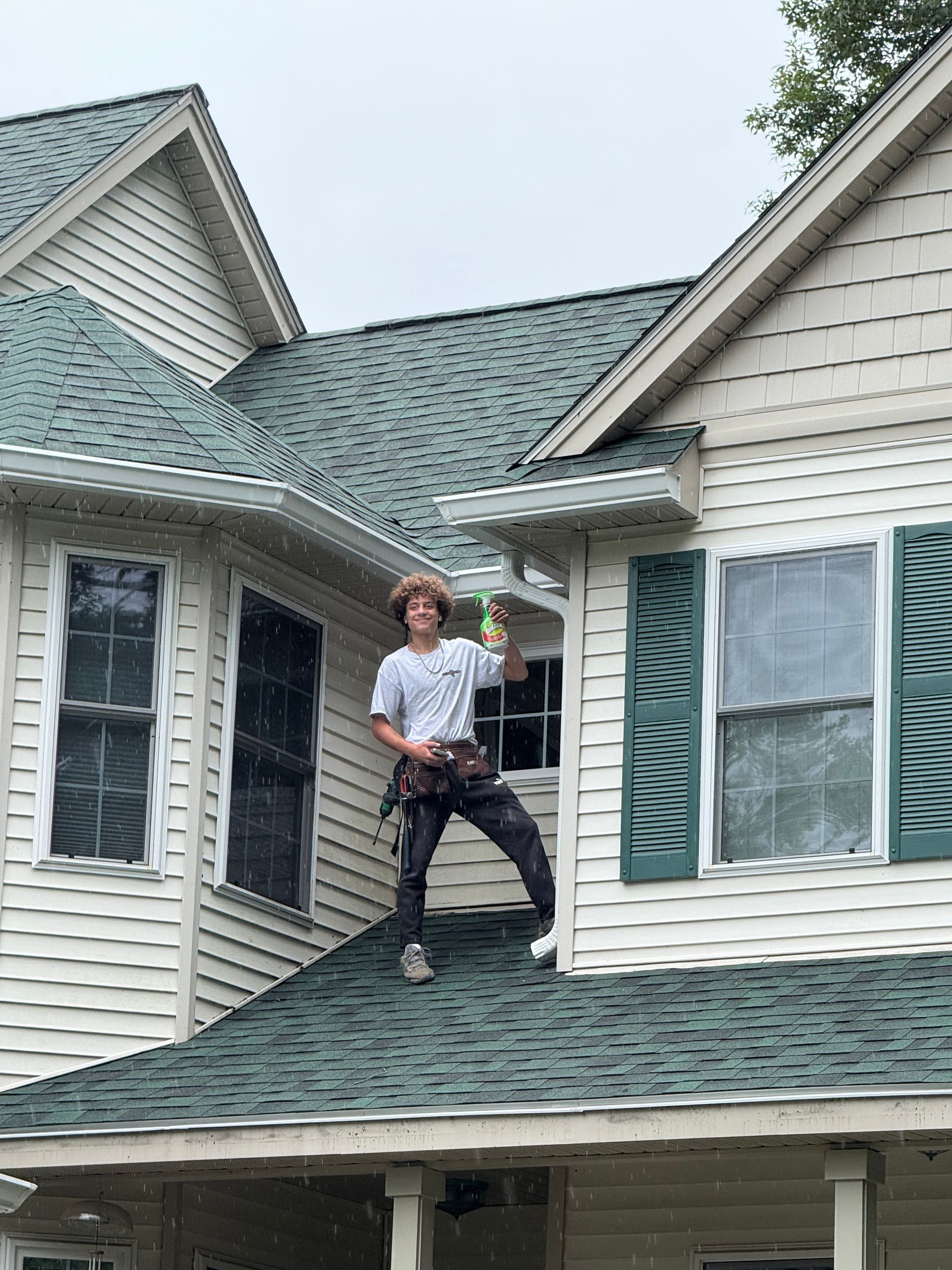 Person standing on a steep house roof, surrounded by light siding and green shutters.