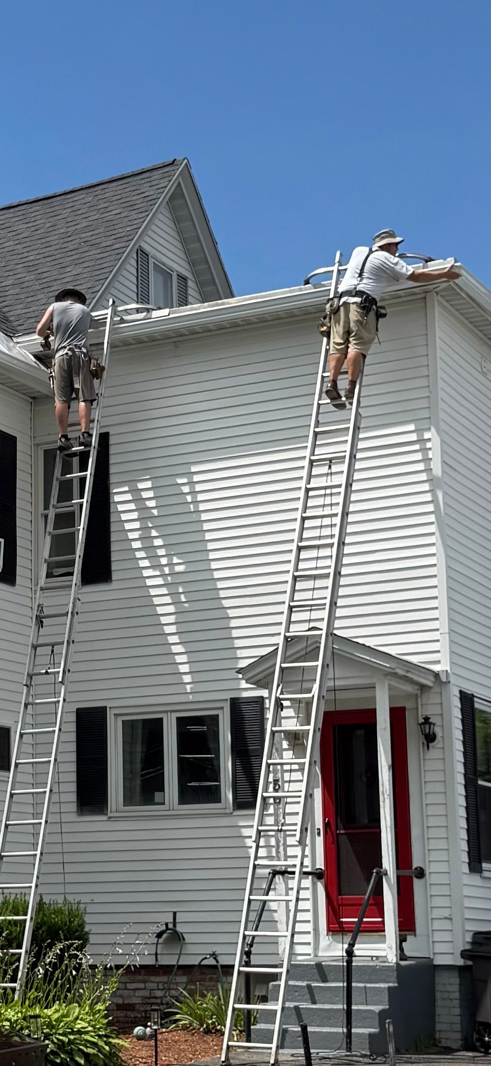 Two workers on ladders painting a white house with black shutters and a red door