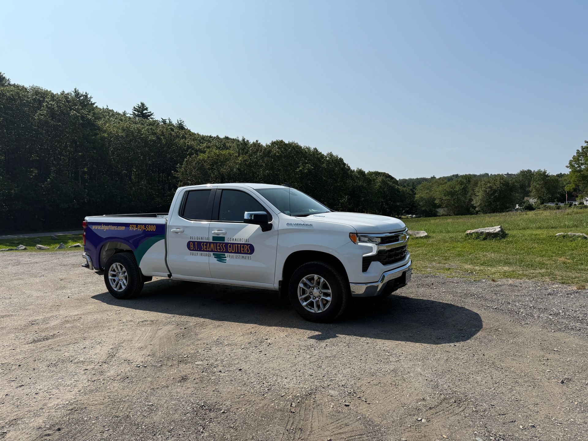 White pickup truck parked on a gravel lot beside a grassy field and trees under a blue sky