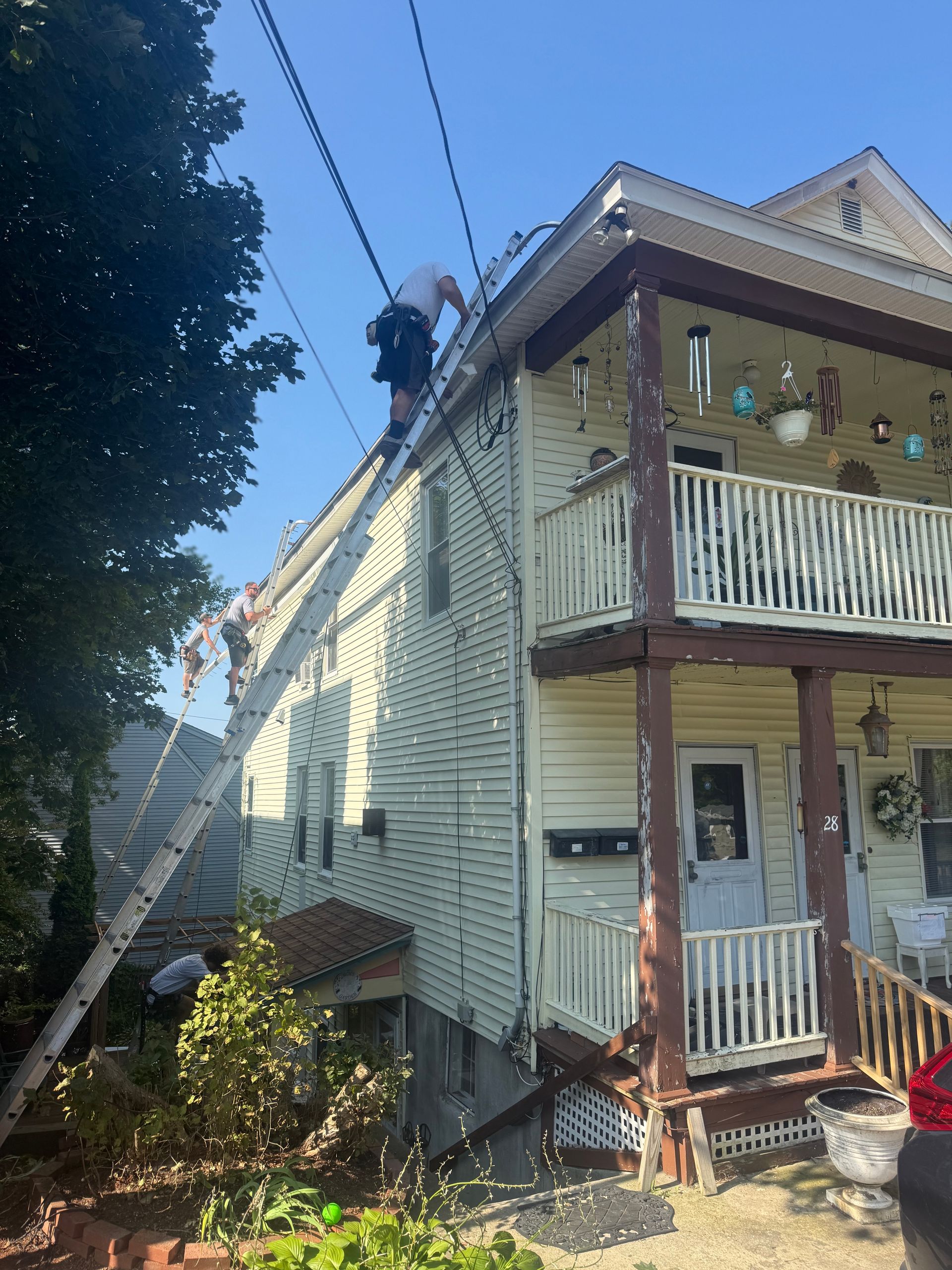 Worker on a ladder repairing the roof of a cream two-story house with a porch and balcony.